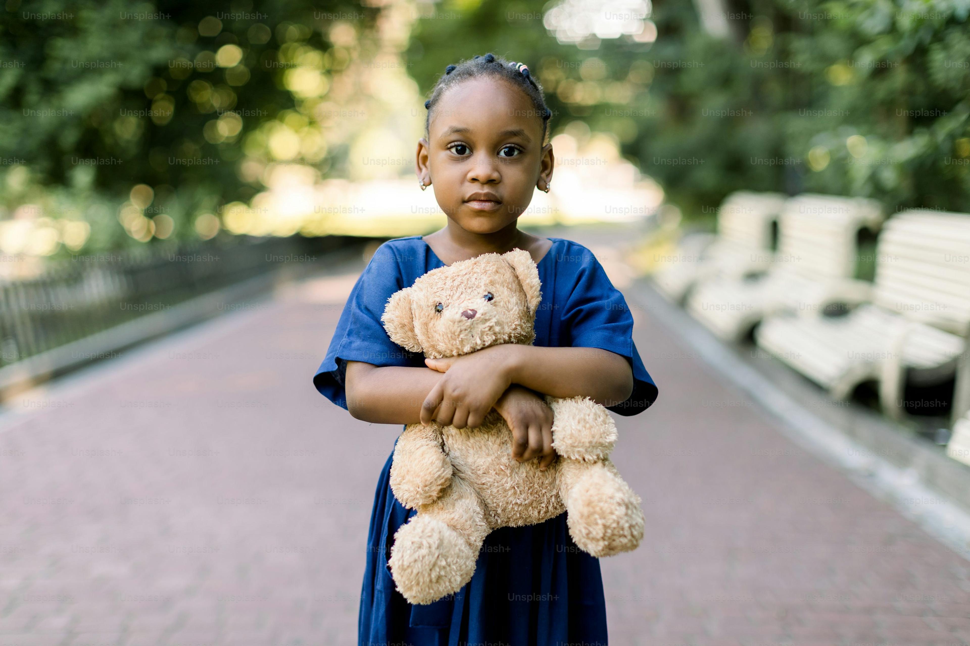 Niña negra sosteniendo un oso de peluche mientras está sola en un camino del parque.