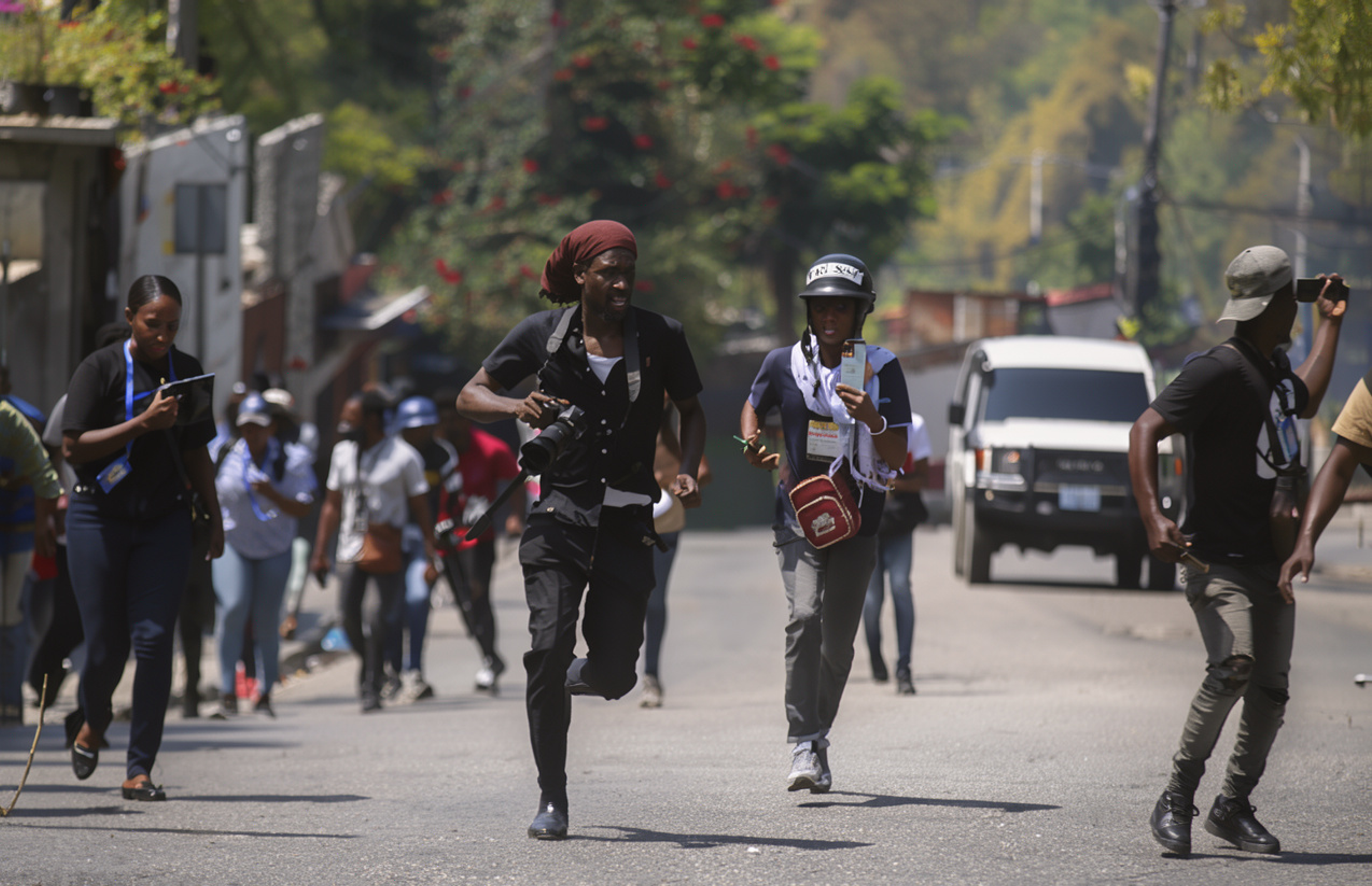 Journalists with cameras and phones run down a street in Haiti, one wearing a “PRESS” helmet.