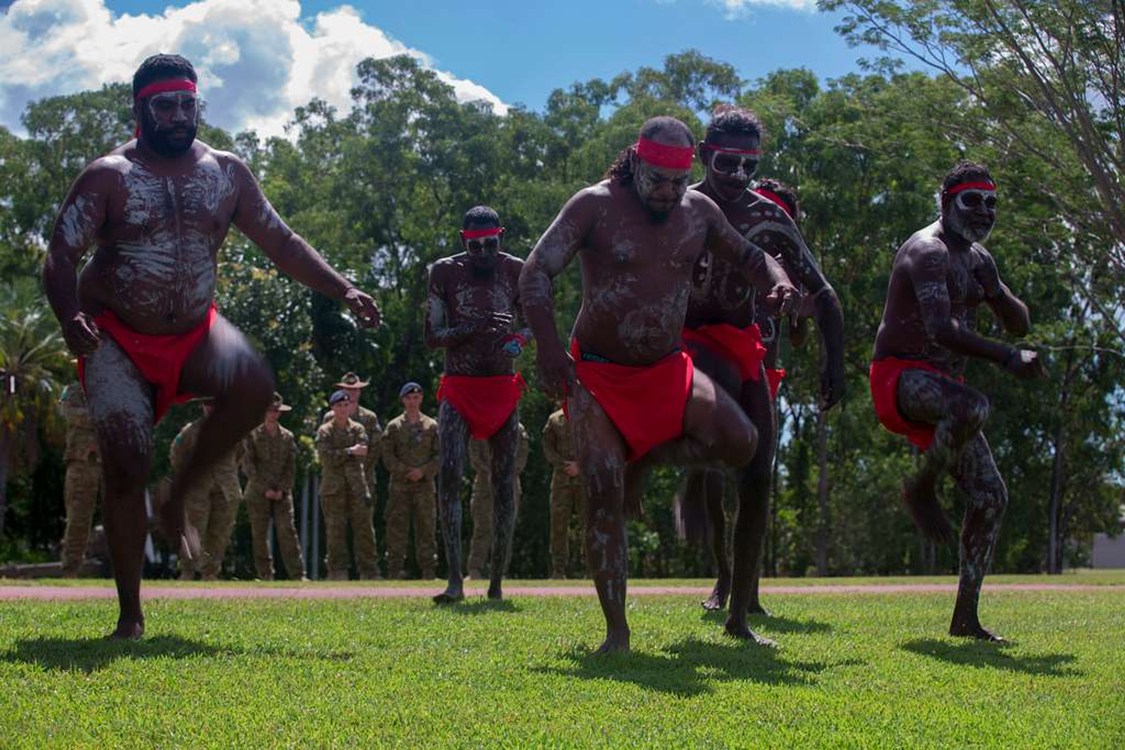 Hombres indígenas realizando un entrenamiento cultural tradicional o danza al aire libre, con soldados observando en el fondo.