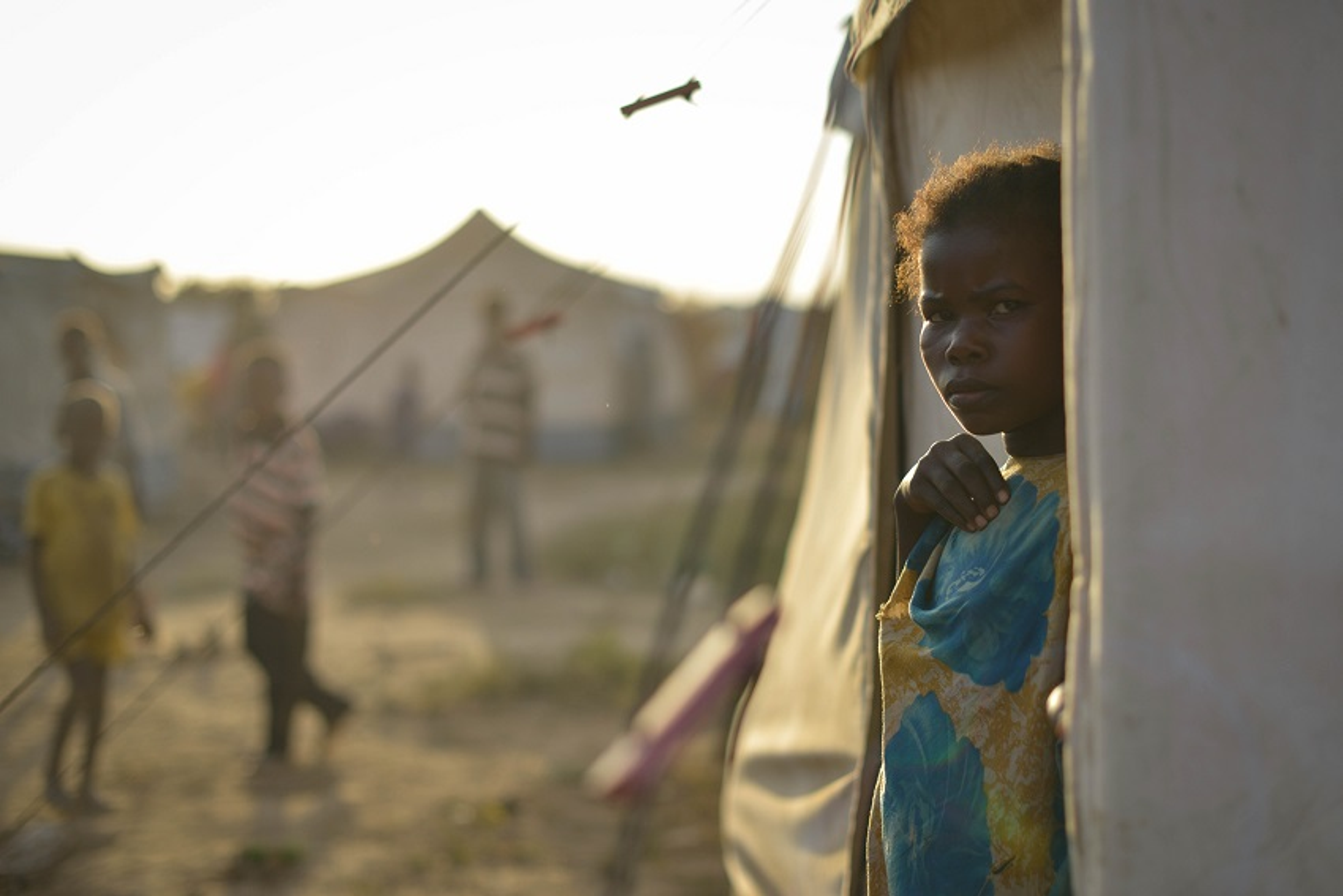 Niña pequeña de pie en la entrada de una tienda en un campamento de refugiados, mirando hacia afuera mientras otros niños caminan en el fondo.