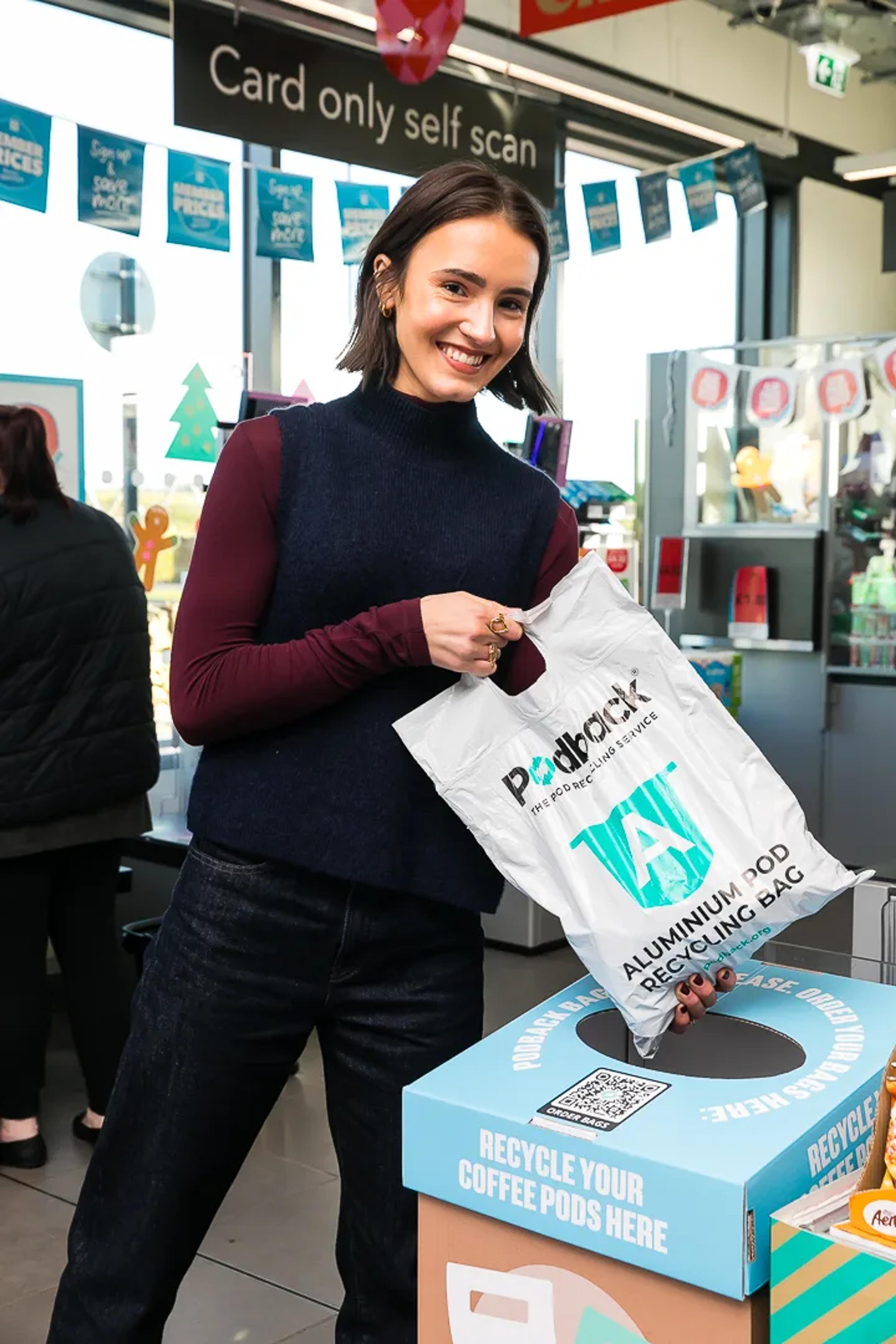 A smiling shopper posting a white Podback aluminium pod recycling bag into a Podback collection box in a supermarket