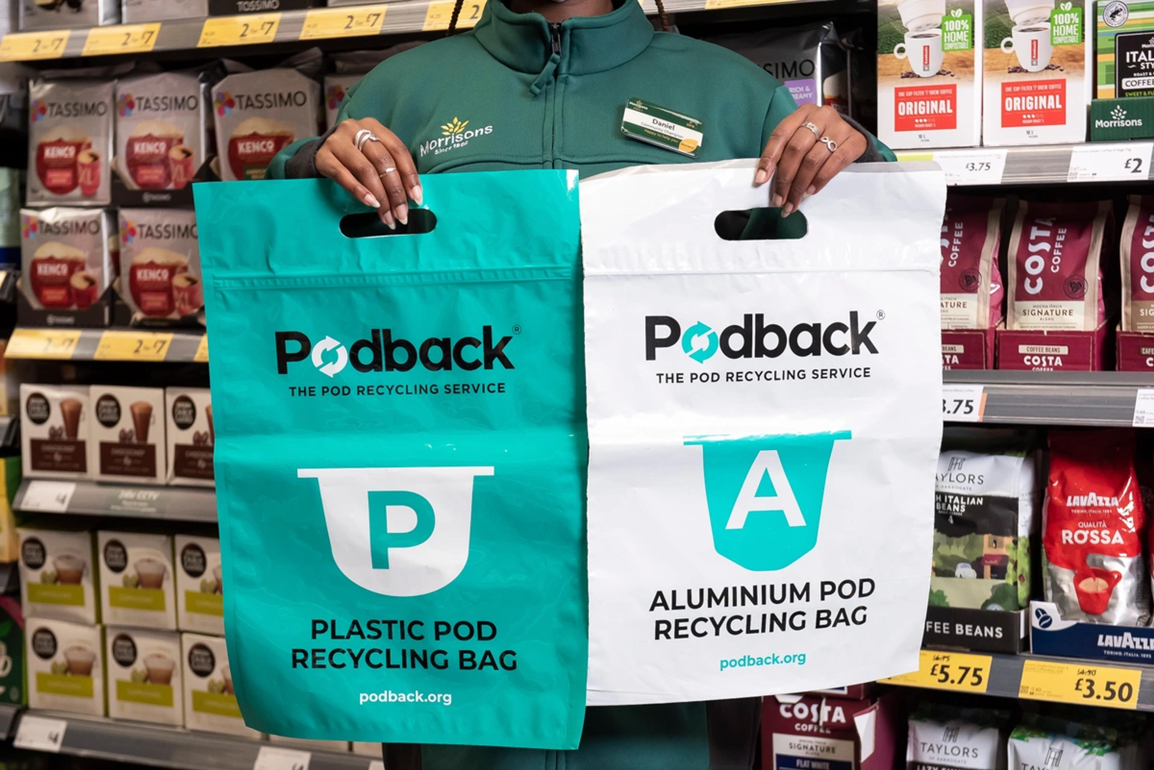 A Morrisons colleague in a supermarket coffee aisle holding up a green Podback plastic pod recycling bag and a white aluminium pod recycling bag.