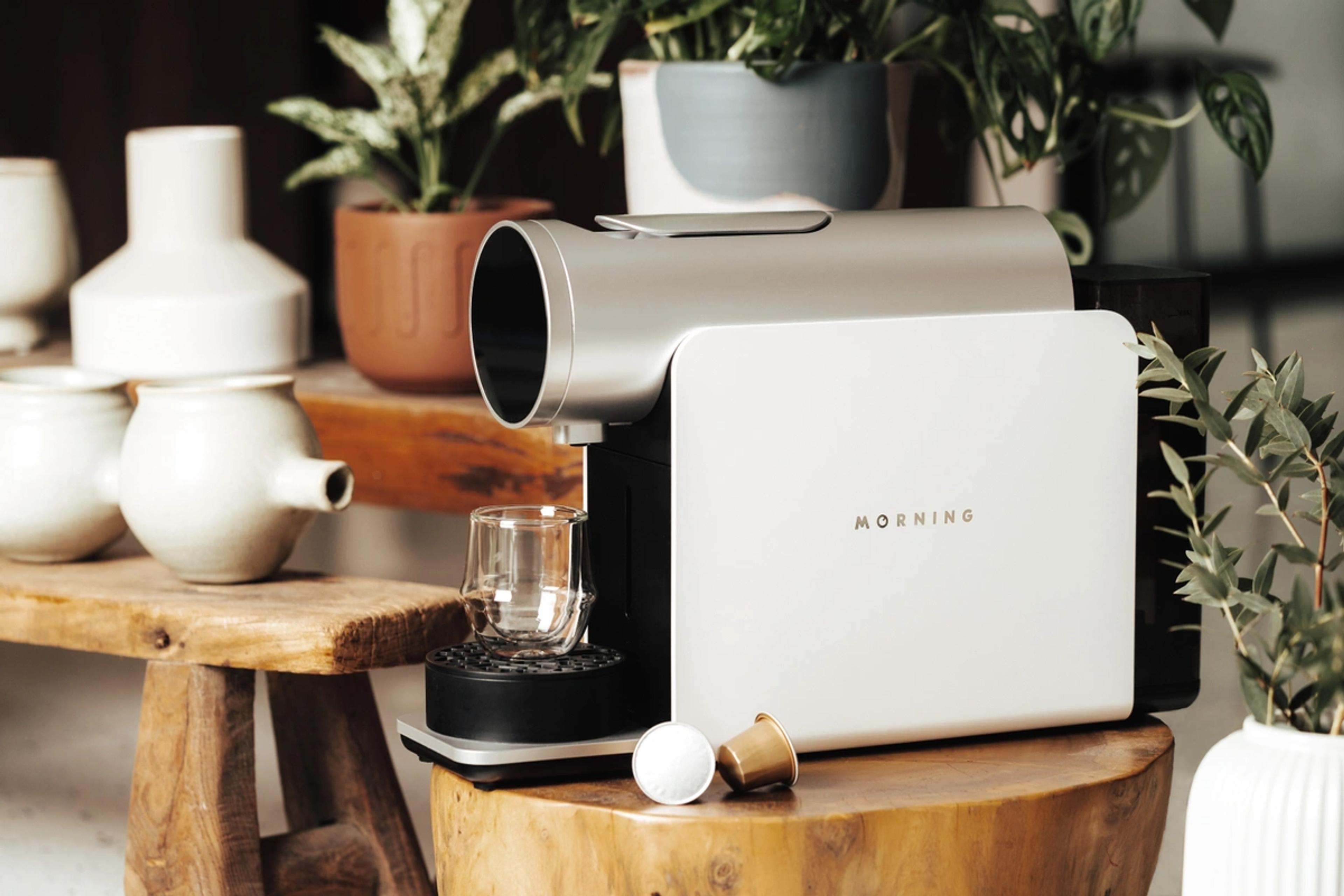 A MORNING coffee pod machine on a wooden stool with a glass cup and two coffee capsules, surrounded by house plants and ceramics.