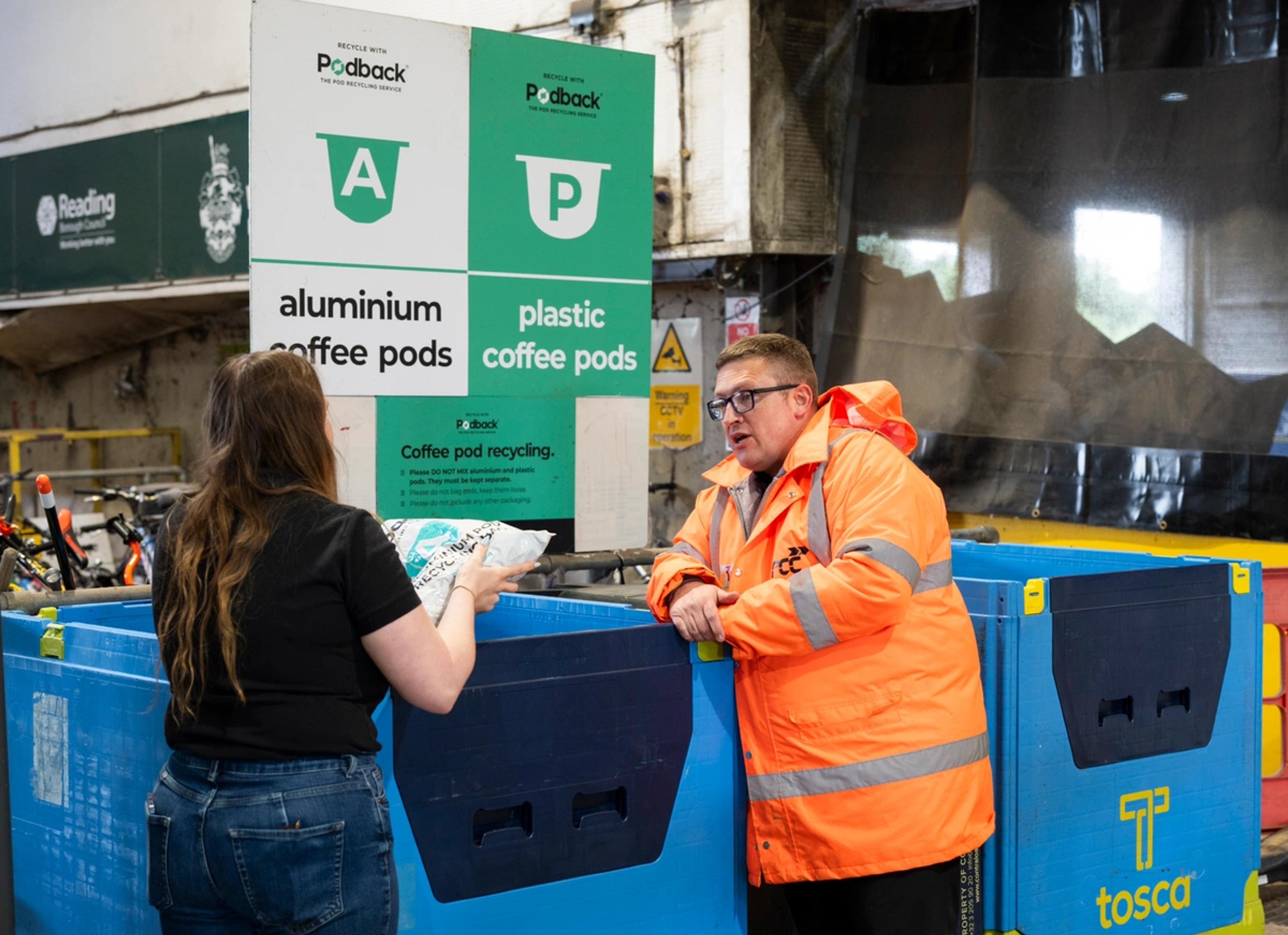 A resident with a bag of coffee pods talks to a site worker in hi-vis beside Podback aluminium and plastic pod containers at a Reading recycling centre.