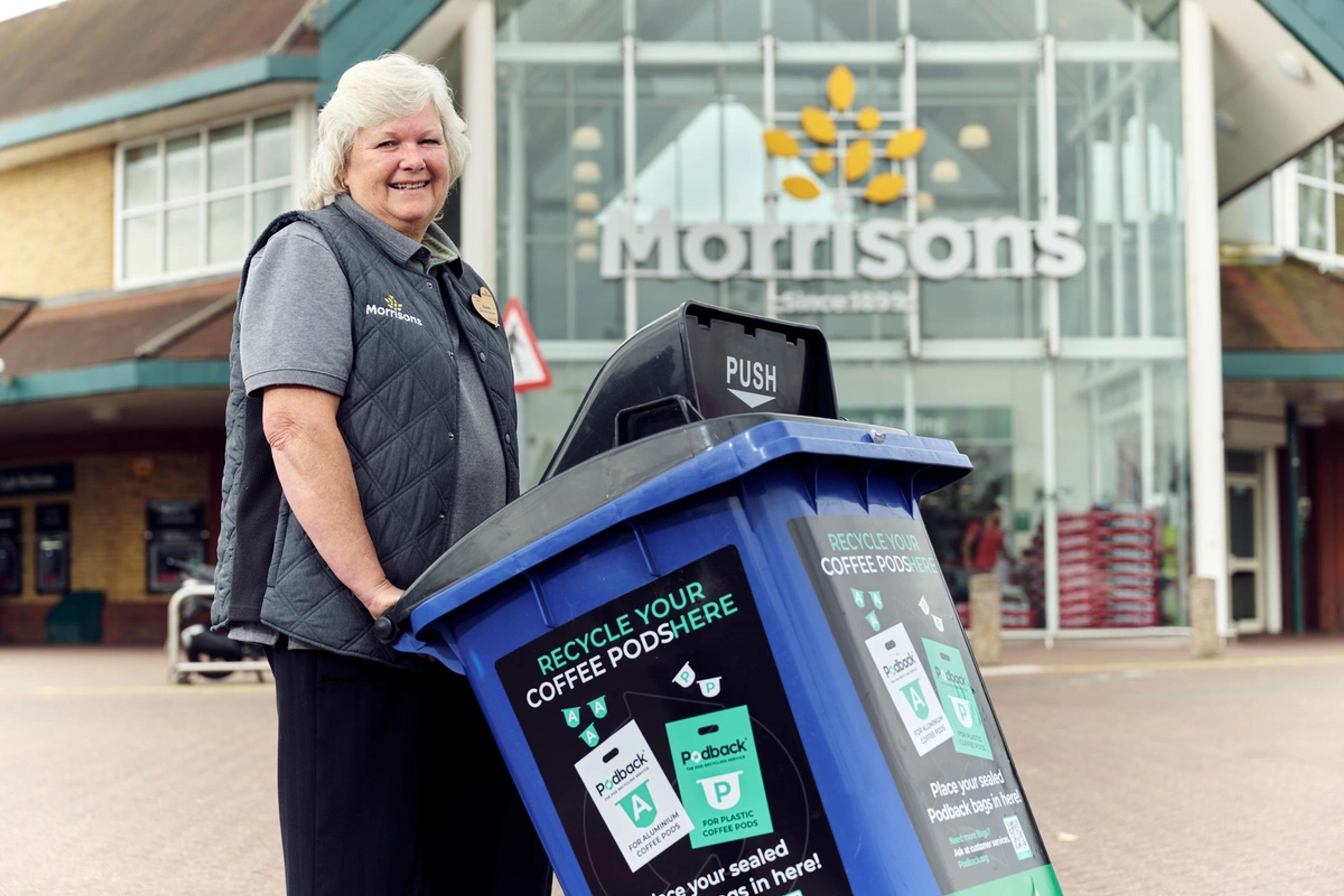 A smiling Morrisons colleague outside a Morrisons store with a blue wheelie bin labelled "Recycle your coffee pods here" for sealed Podback bags.