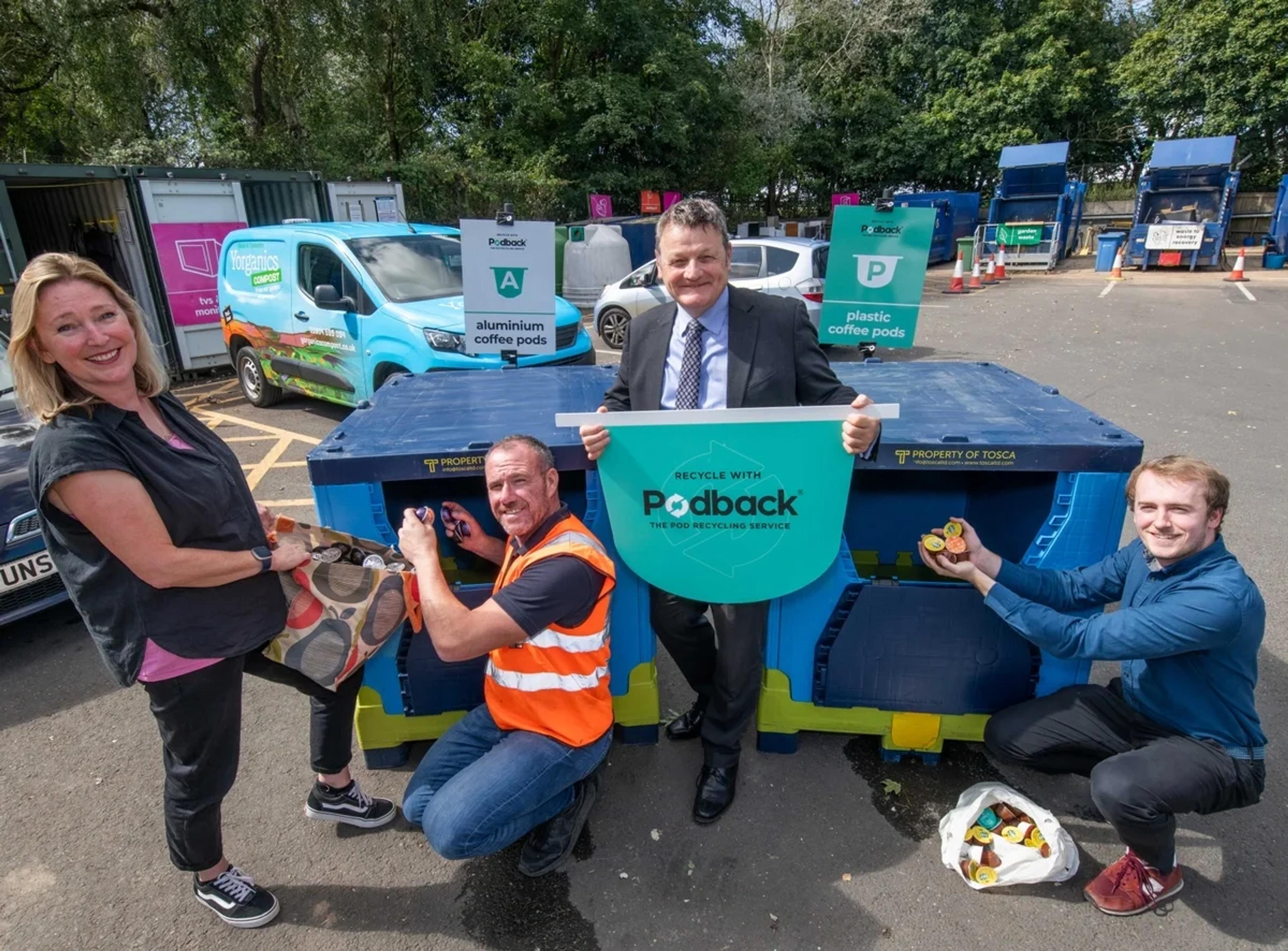 Four people at a recycling centre with blue Podback containers for aluminium and plastic coffee pods, one holding a "Recycle with Podback" sign and others dropping pods in.