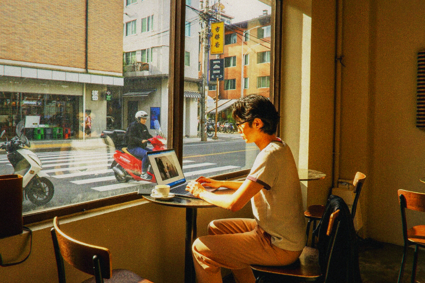 Young man with glasses working on a laptop by a cafe window as traffic passes outside