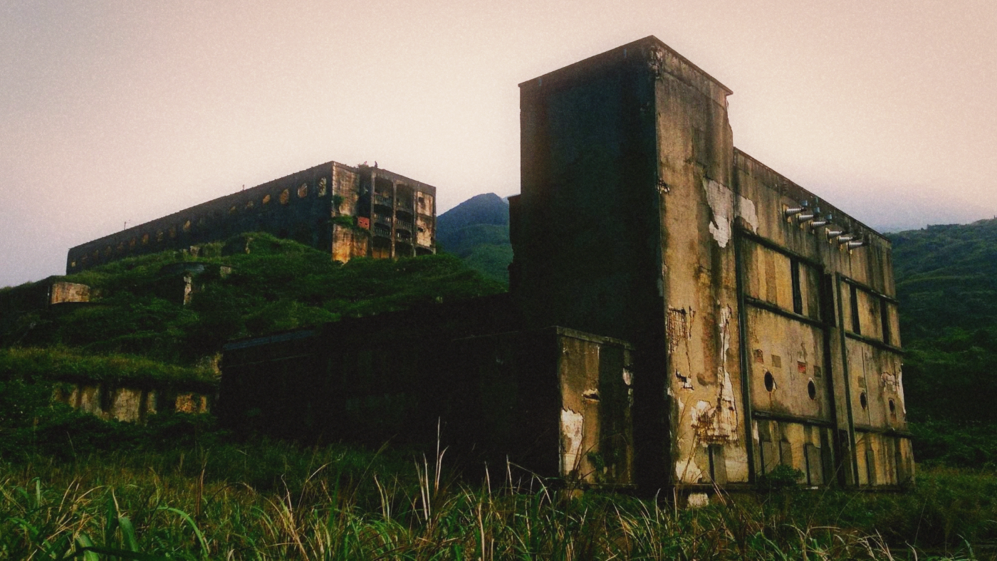 Overgrown concrete ruins of a terraced smelter standing in tall grass below hills