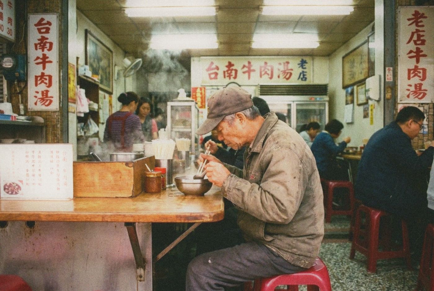 Side view of a customer sitting on a plastic stool eating beef soup at a counter in a traditional Tainan shop