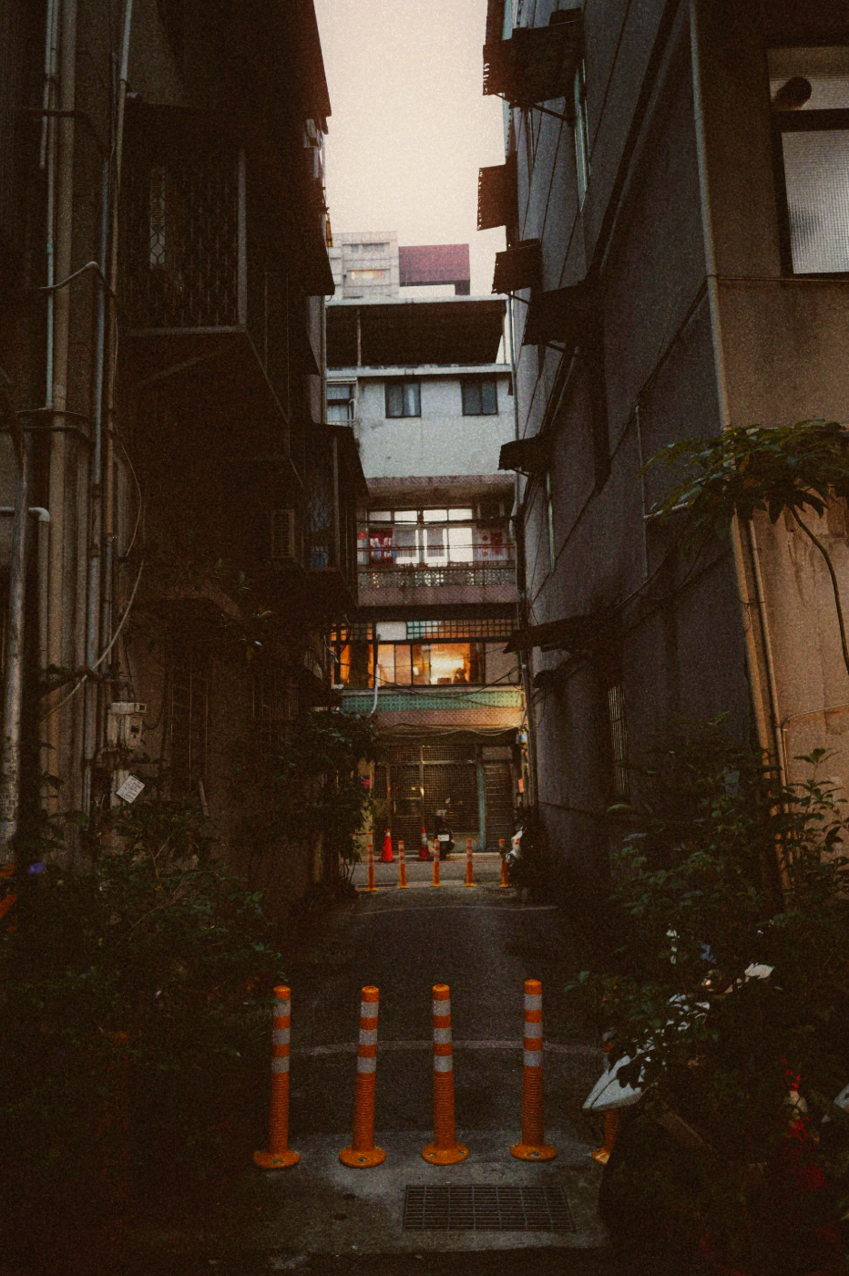 Narrow alley between weathered apartment buildings at dusk, orange bollards in the foreground