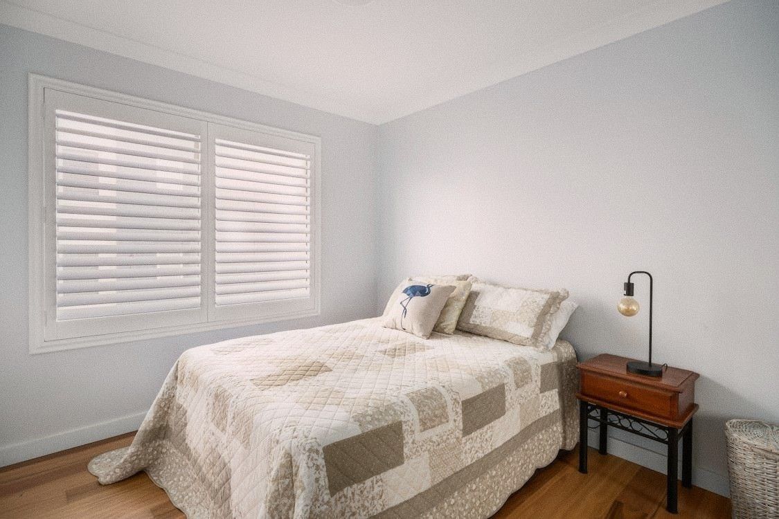 A bedroom with light cotton bedding, window slightly open, minimal furniture and one dim lamp