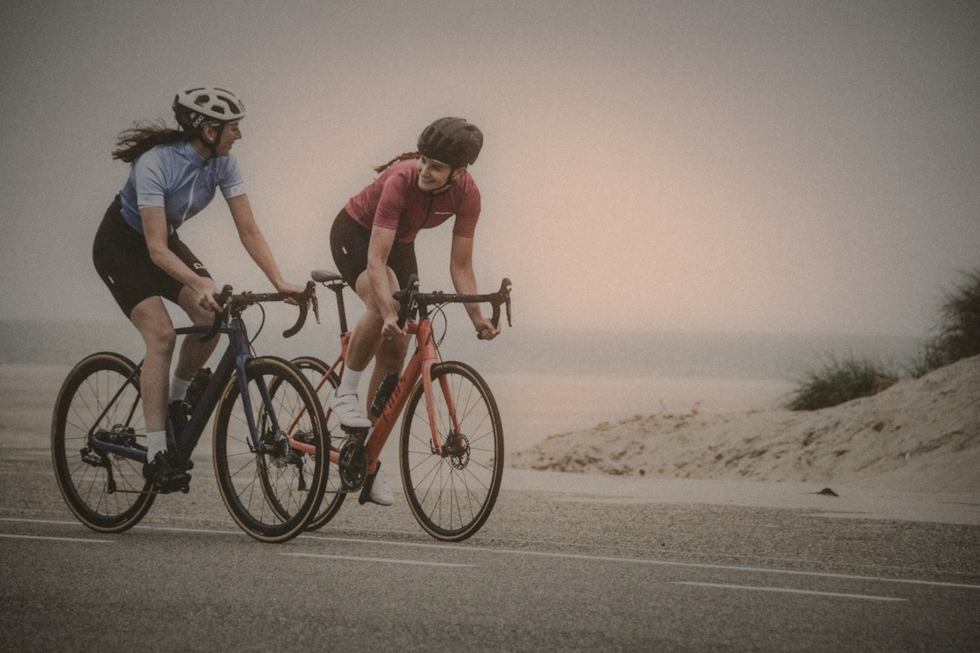 Two cyclists in jerseys and helmets riding side by side on a road beside sand dunes