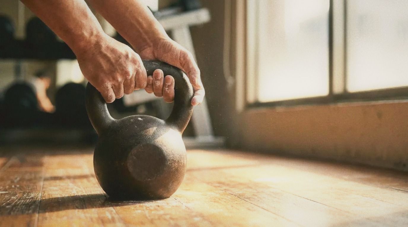 Close-up of hands gripping a kettlebell on a wooden gym floor, chalk visible, warm natural light