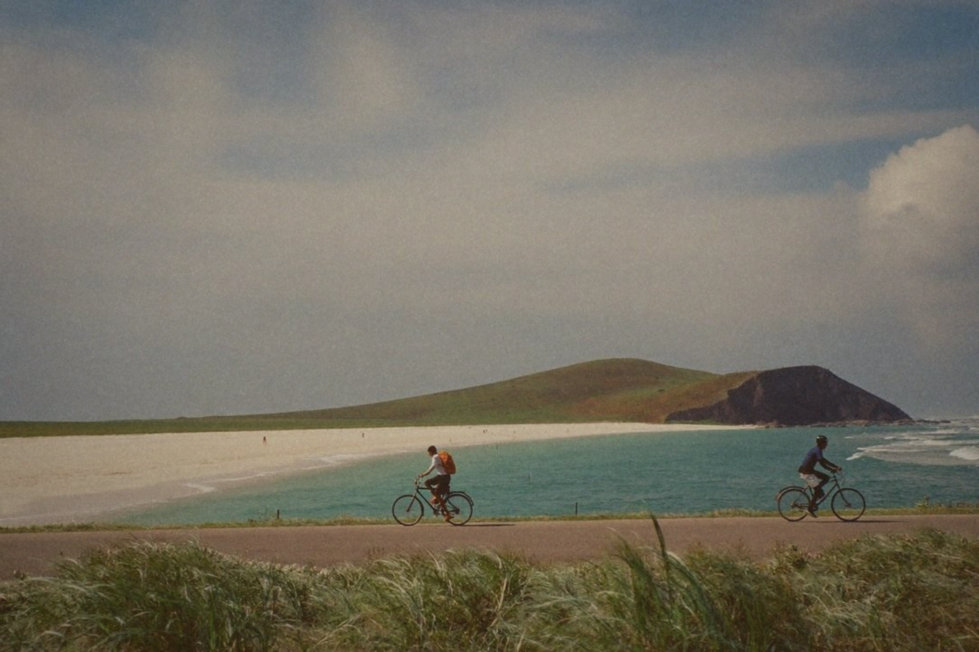 Cyclists riding a flat seaside path on Taiwan's northeast coast beside pale sand and turquoise water with a tunnel mouth in a green headland