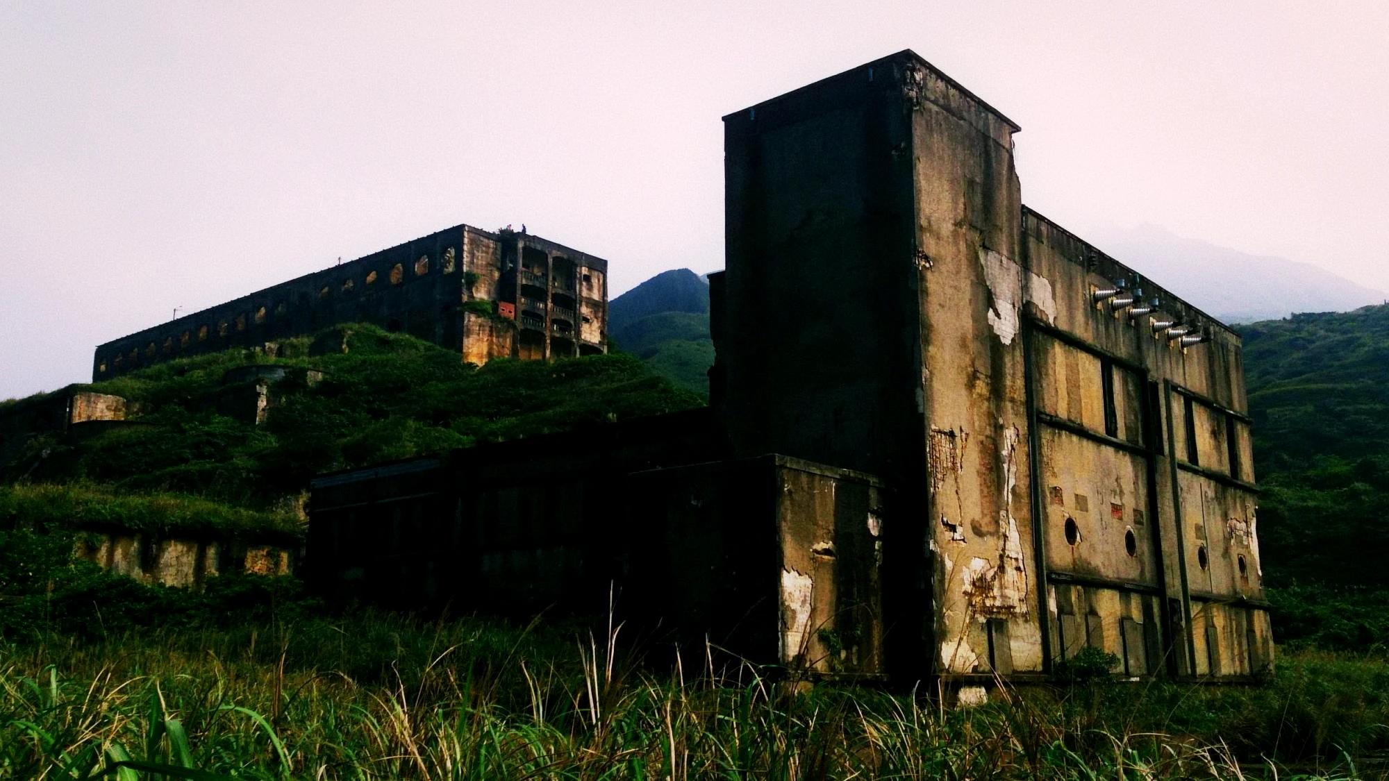 Overgrown concrete ruins of a terraced smelter standing in tall grass below hills