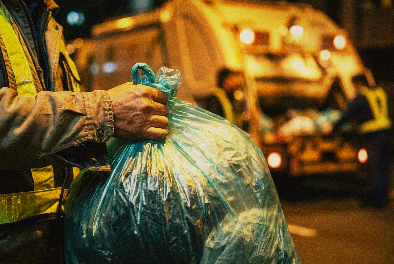 A yellow Taipei garbage truck at dusk with neighbours lining up to hand over blue-green rubbish bags, scooters and apartment buildings behind, warm streetlight
