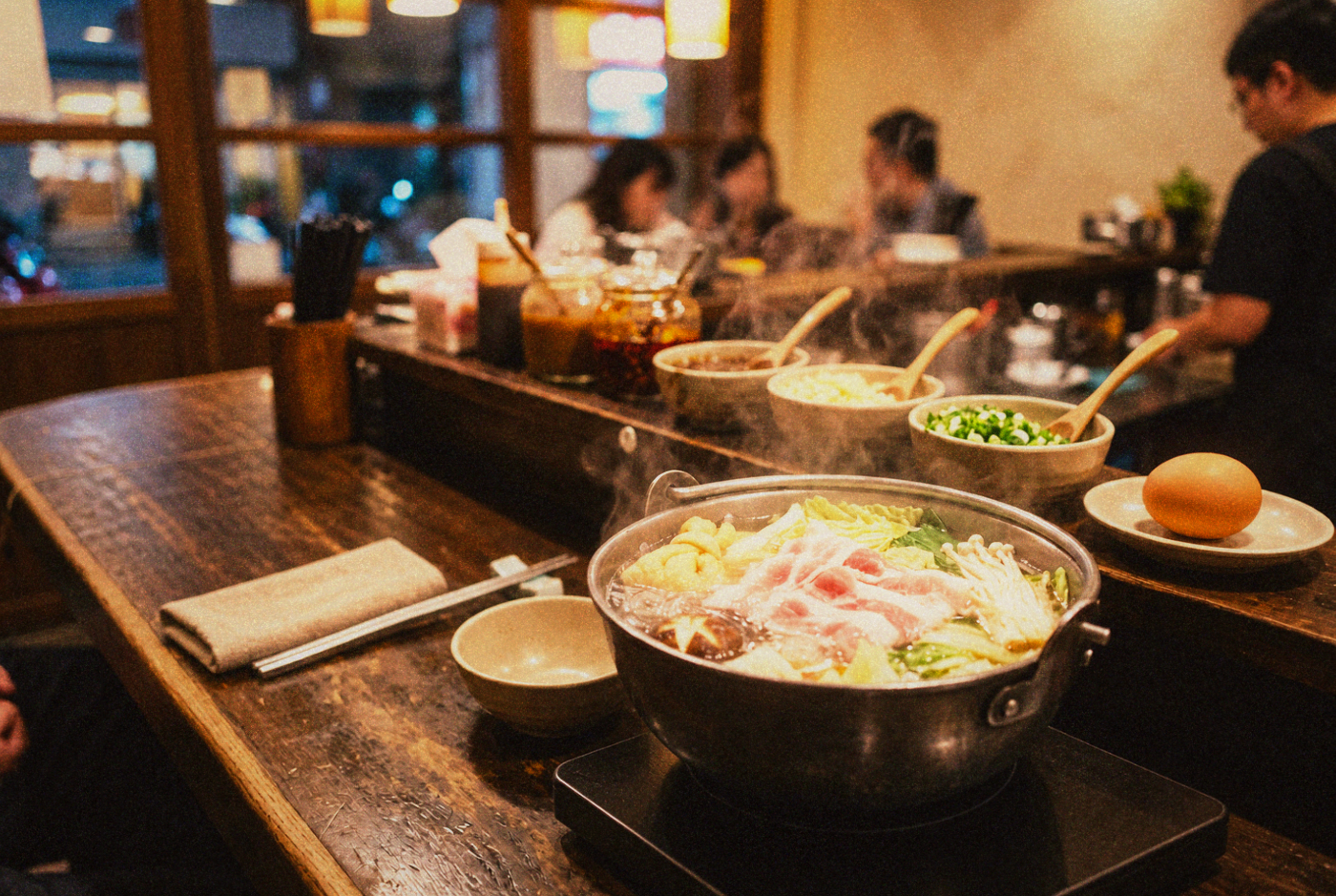 A single personal hot pot simmering on a Taipei restaurant counter, one portion of thin-sliced meat and vegetables, a self-serve sauce station blurred in the background, warm evening light