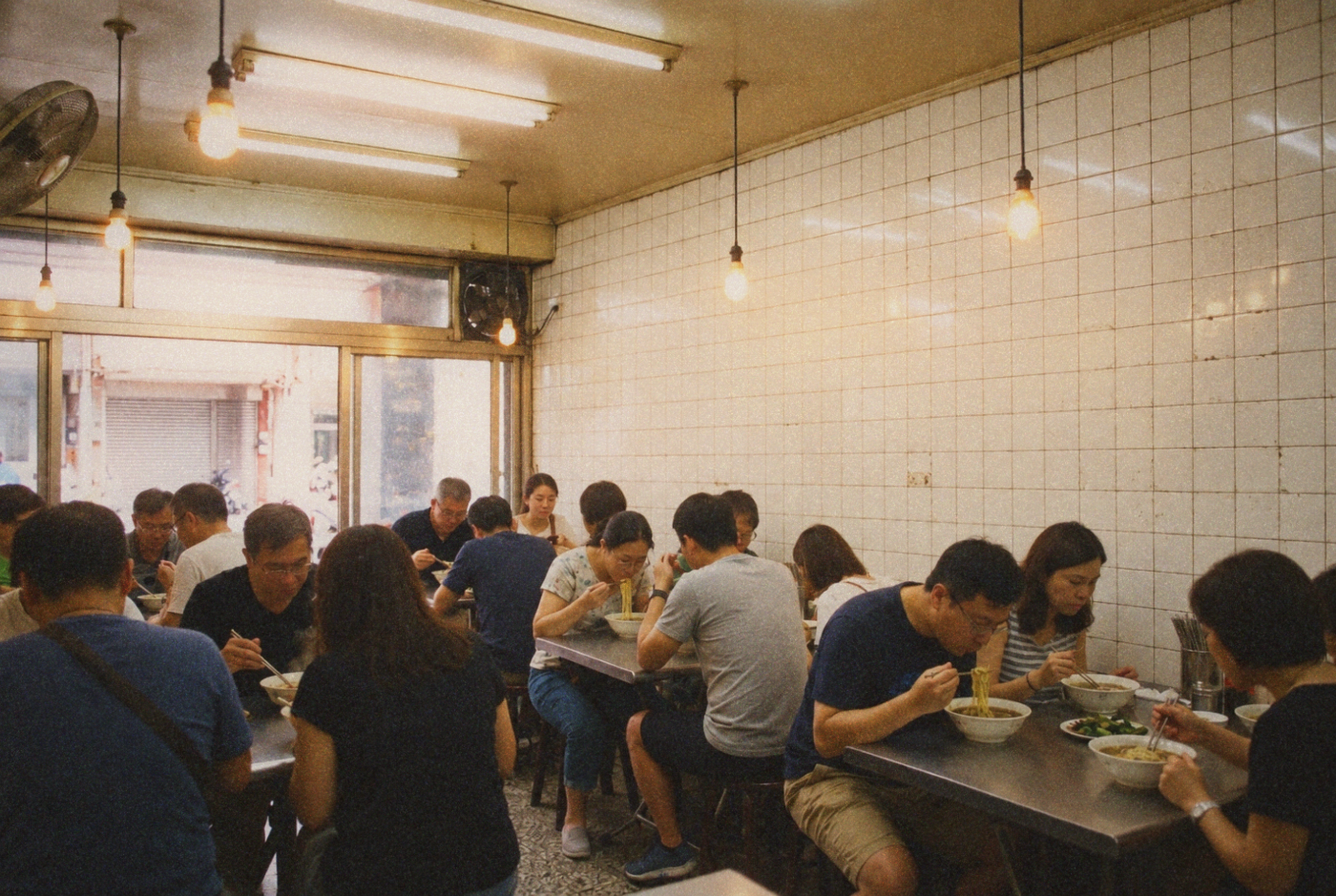 Diners eating noodles at stainless-steel tables in a tiled shop