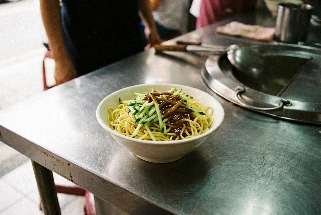 A bowl of Taiwanese cold noodles topped with shredded cucumber and sesame sauce on a shop counter