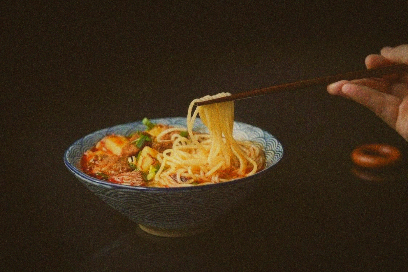 Hand lifting thin noodles with chopsticks from a patterned bowl of red-brown broth