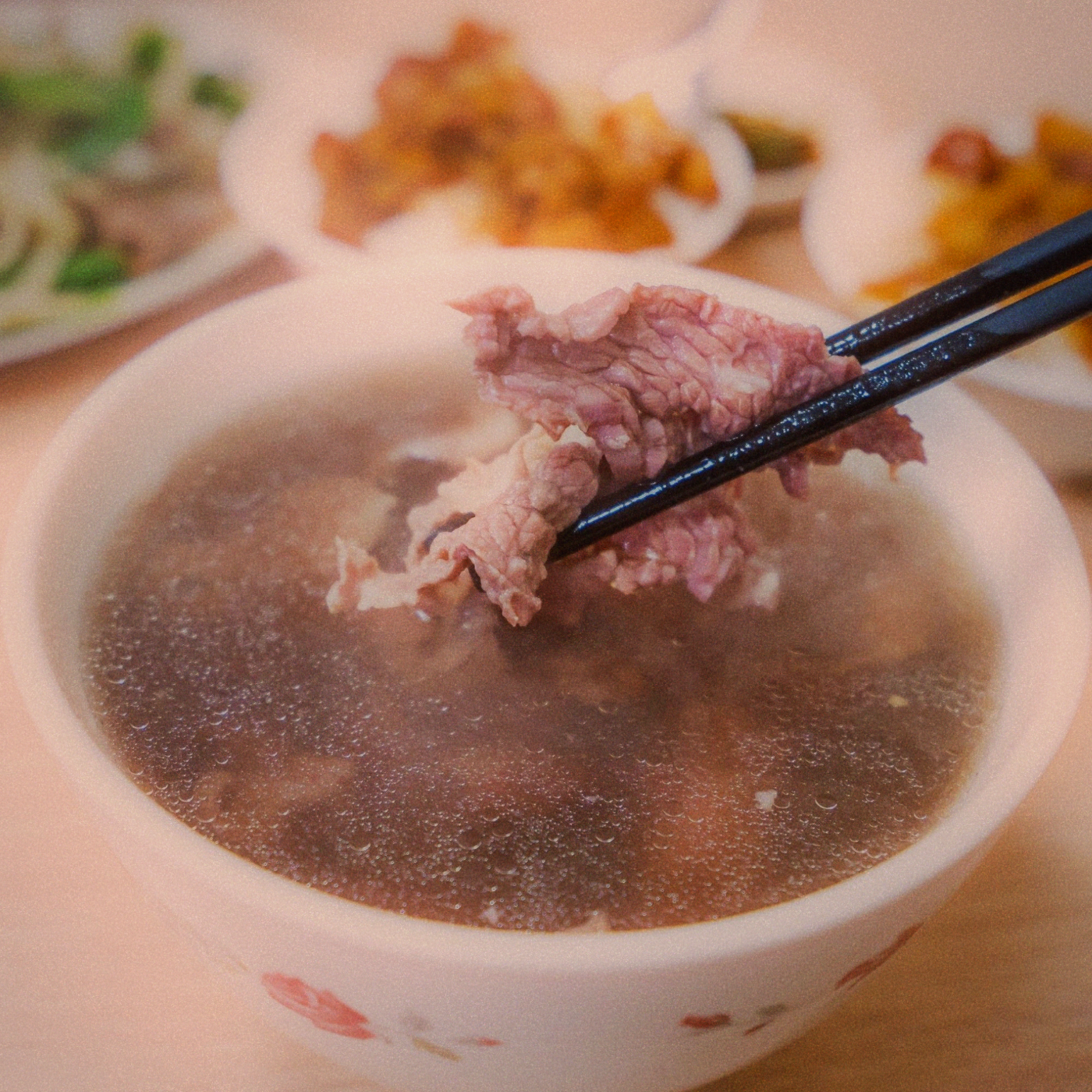 Chopsticks lifting a slice of rare beef from a bowl of clear beef soup