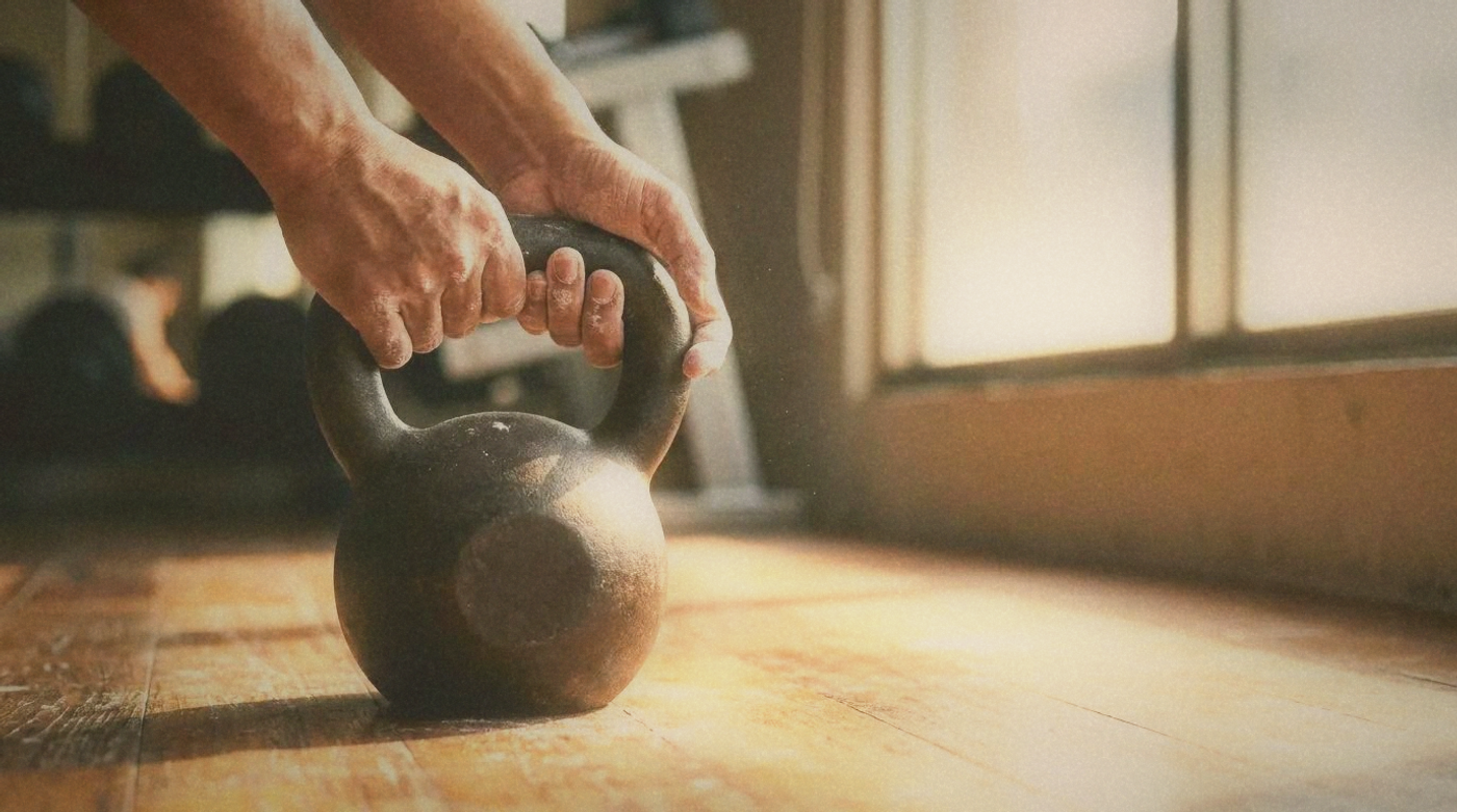 Close-up of hands gripping a kettlebell on a wooden gym floor, chalk visible, warm natural light