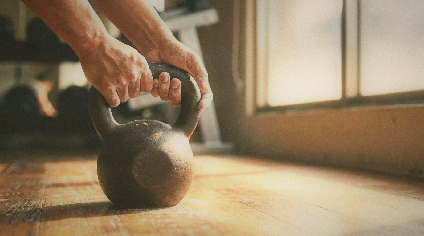 Close-up of hands gripping a kettlebell on a wooden gym floor, chalk visible, warm natural light