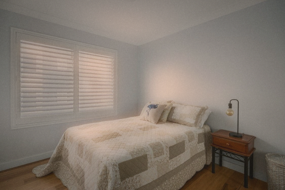 A bedroom with light cotton bedding, window slightly open, minimal furniture and one dim lamp
