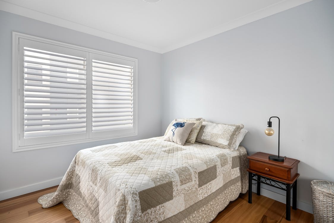 A bedroom with light cotton bedding, window slightly open, minimal furniture and one dim lamp