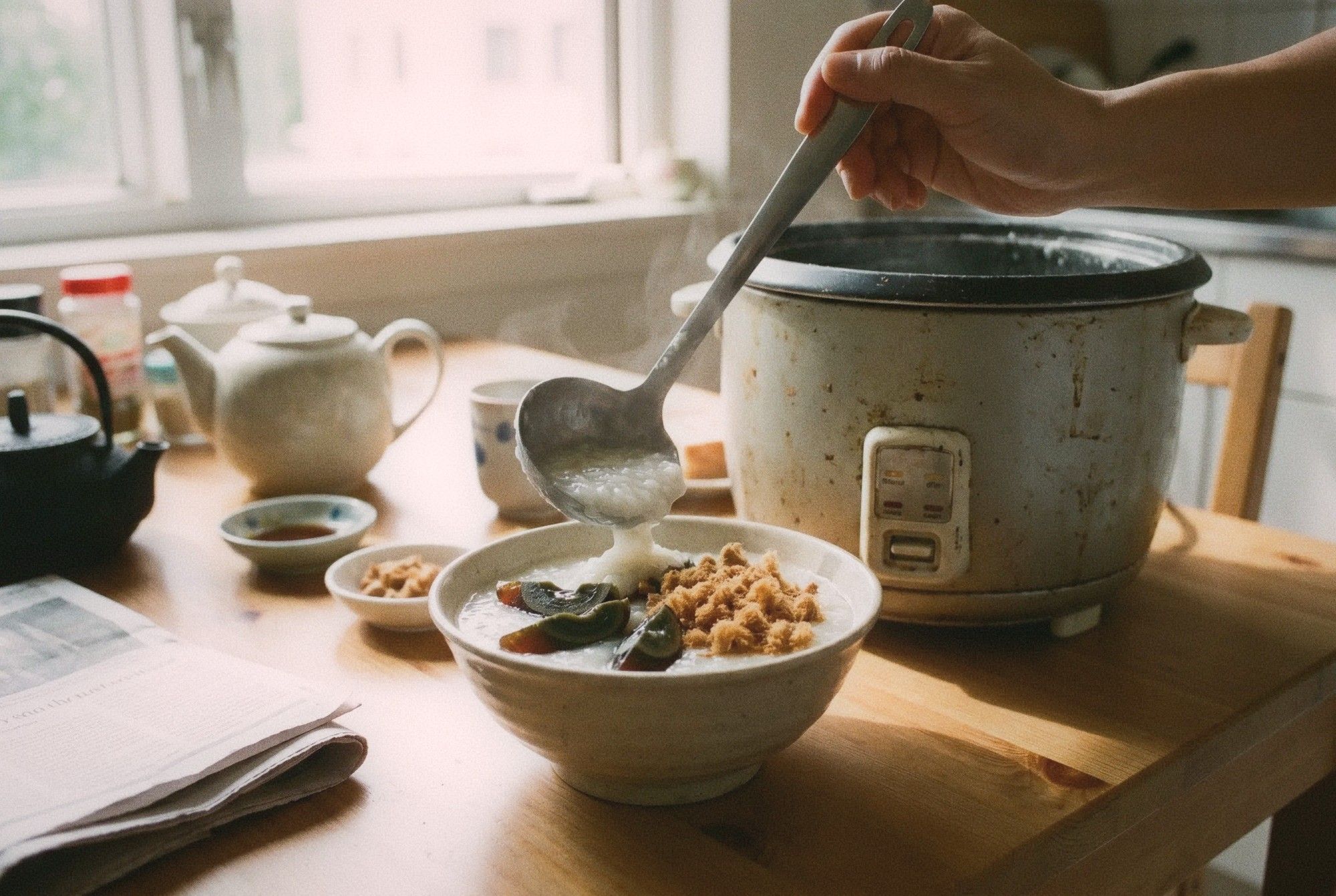 Bowl of congee rice porridge with century egg and pork floss toppings being ladled from a rice cooker