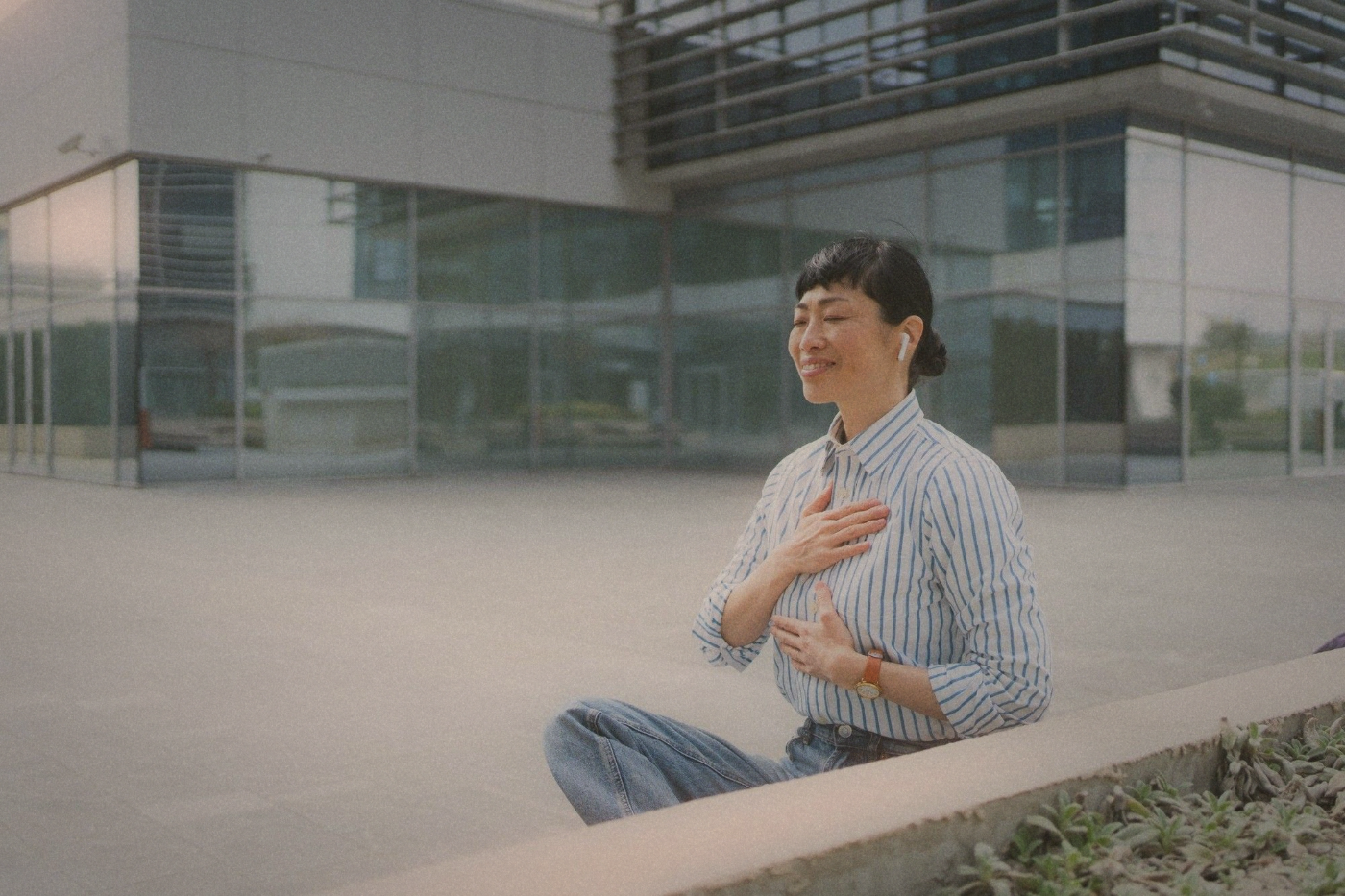 Woman sitting cross-legged on a low wall outside a glass office building, hand resting on her chest