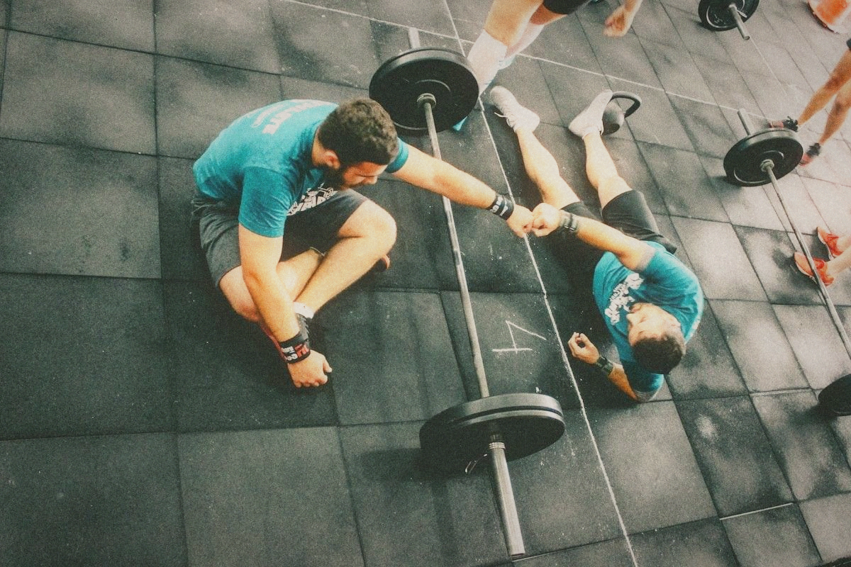 Two men in matching shirts bumping fists on a gym floor beside loaded barbells
