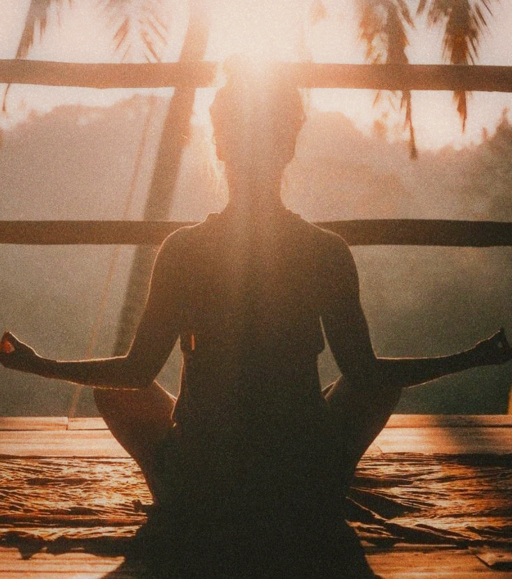 Silhouette of a person sitting cross-legged on a wooden deck, backlit by sun through palm trees