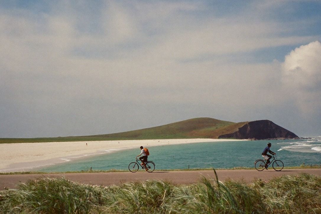 Cyclists riding a flat seaside path on Taiwan's northeast coast beside pale sand and turquoise water with a tunnel mouth in a green headland