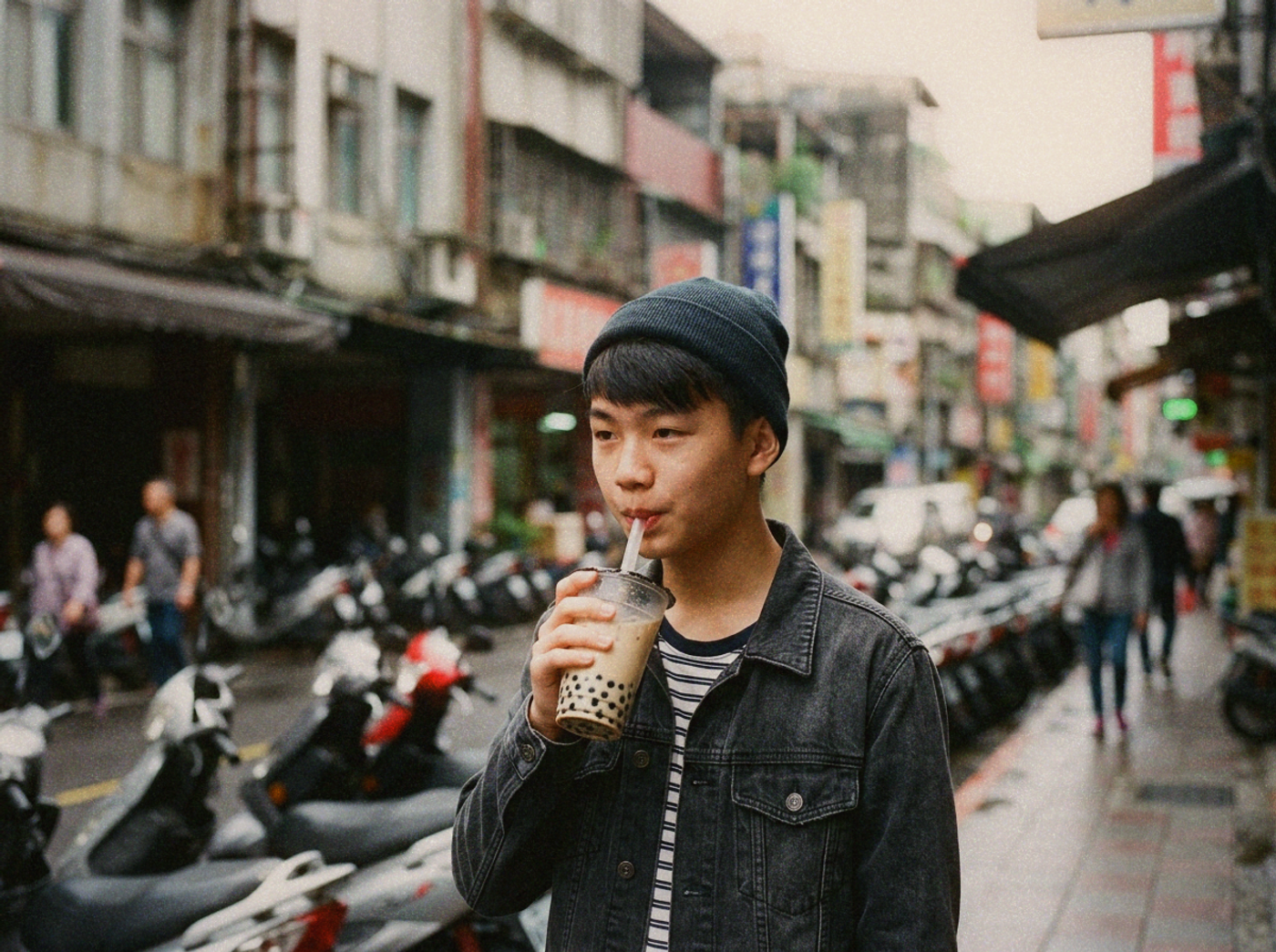 A young person on a Taipei street mid-sip holding a sealed cup of bubble tea with scooters blurred behind