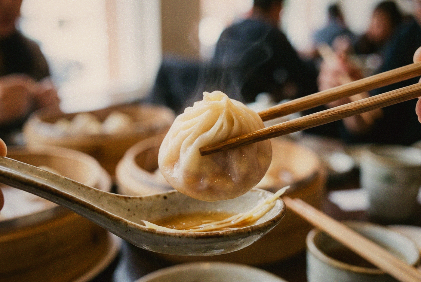 A single translucent Taiwanese xiaolongbao soup dumpling lifted on chopsticks over a spoon, thin skin glistening with broth inside