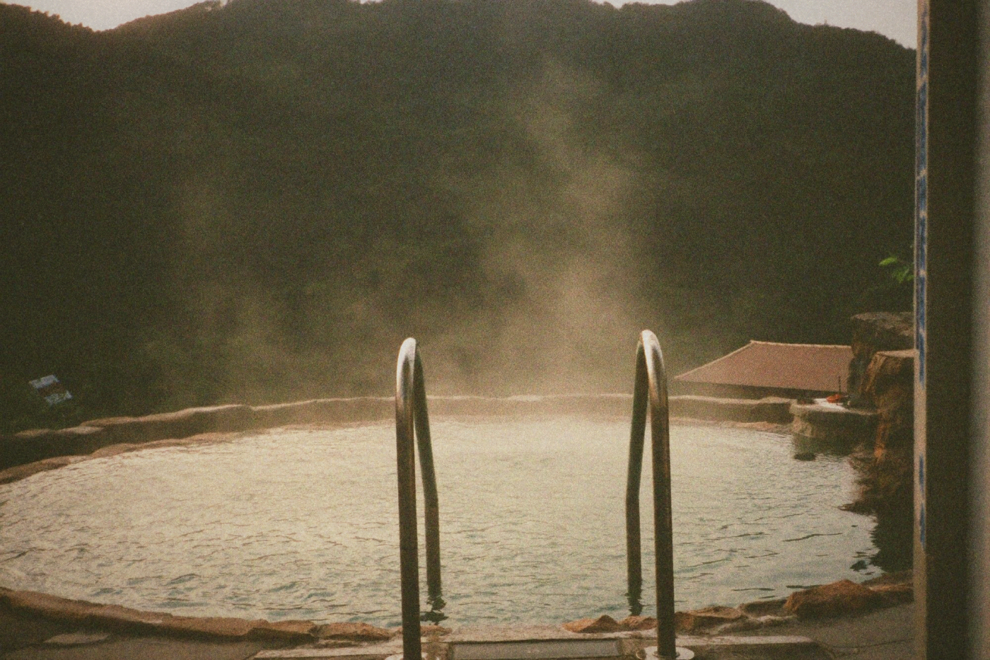 Metal handrail descending into a steaming hot spring pool with a small shelter nearby