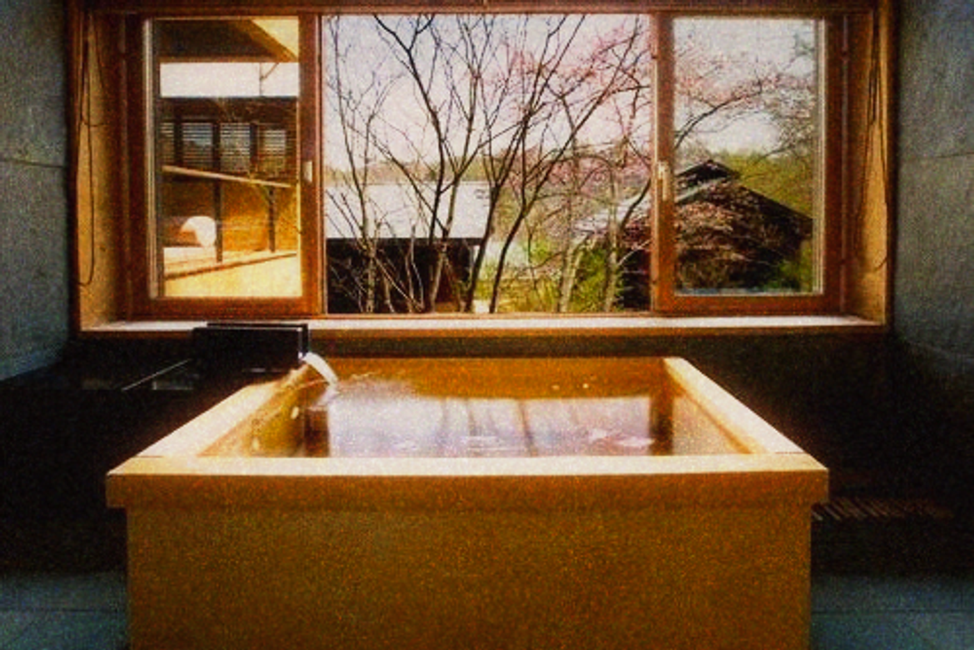 Square wooden soaking tub filled with water in front of a window looking onto bare trees