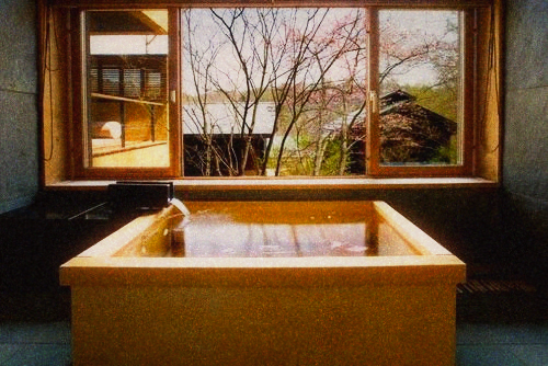 Square wooden soaking tub filled with water in front of a window looking onto bare trees