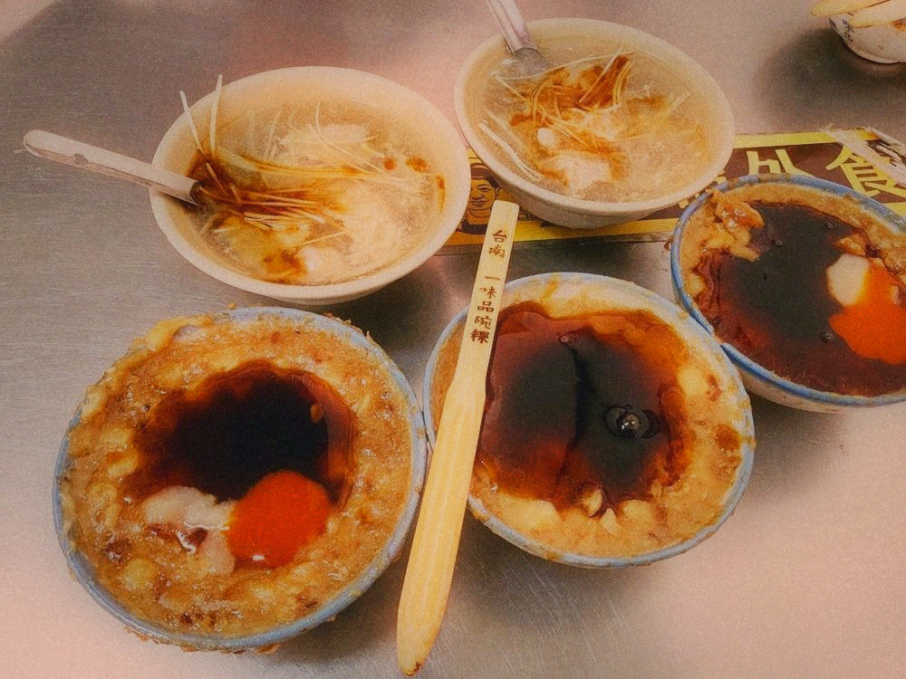 Four bowls of savoury rice pudding topped with dark sauce beside two bowls of clear soup