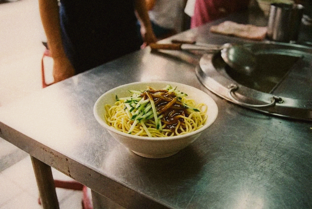 A bowl of Taiwanese cold noodles topped with shredded cucumber and sesame sauce on a shop counter