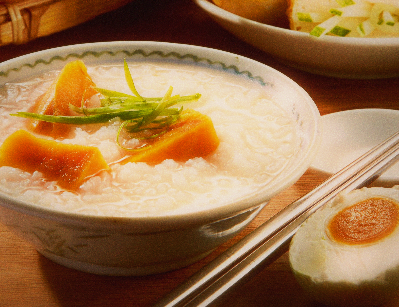 Bowl of rice congee with sweet potato chunks and shredded spring onion beside a halved salted egg