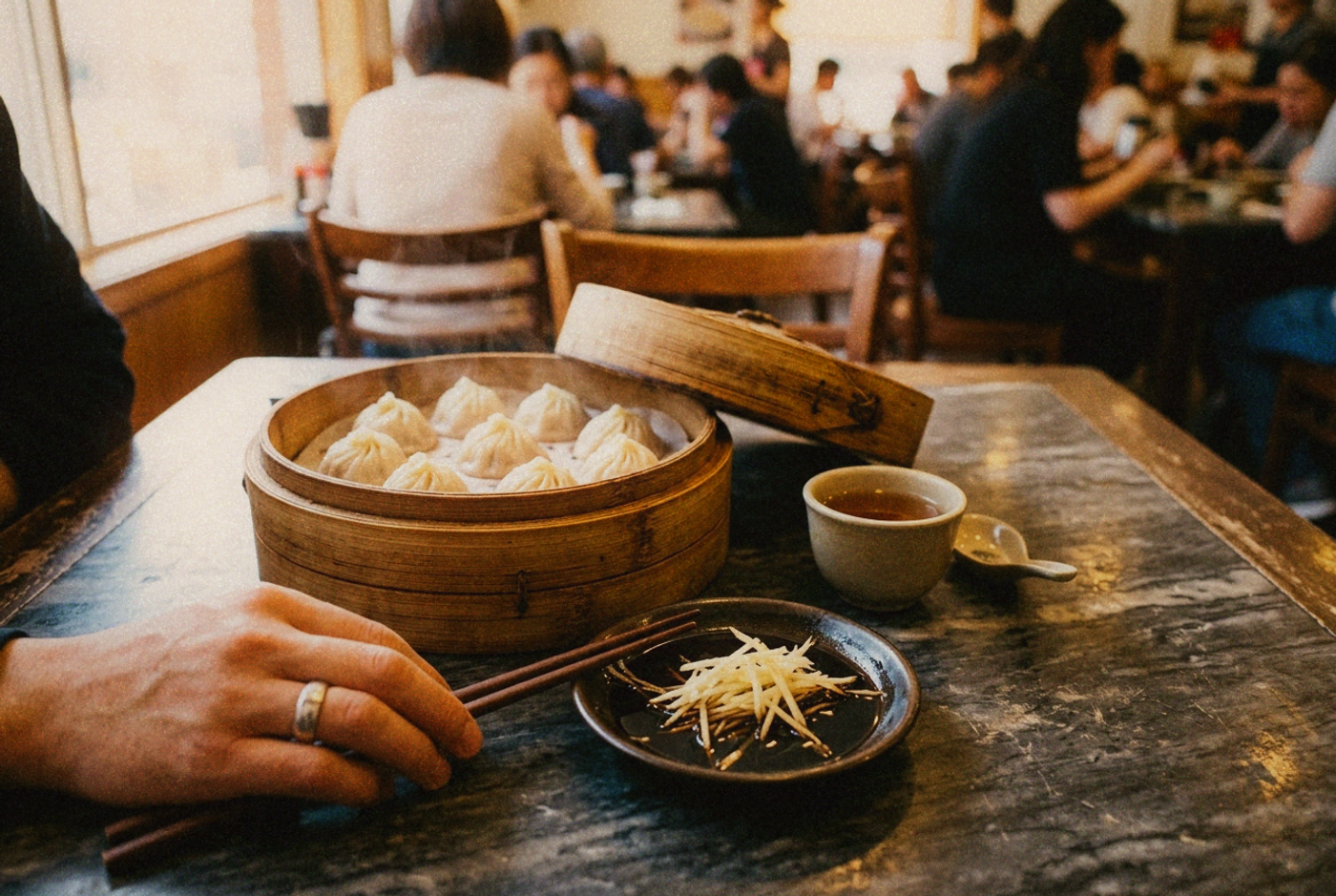 A shallow dish of black vinegar with fine ginger threads beside a bamboo steamer of soup dumplings