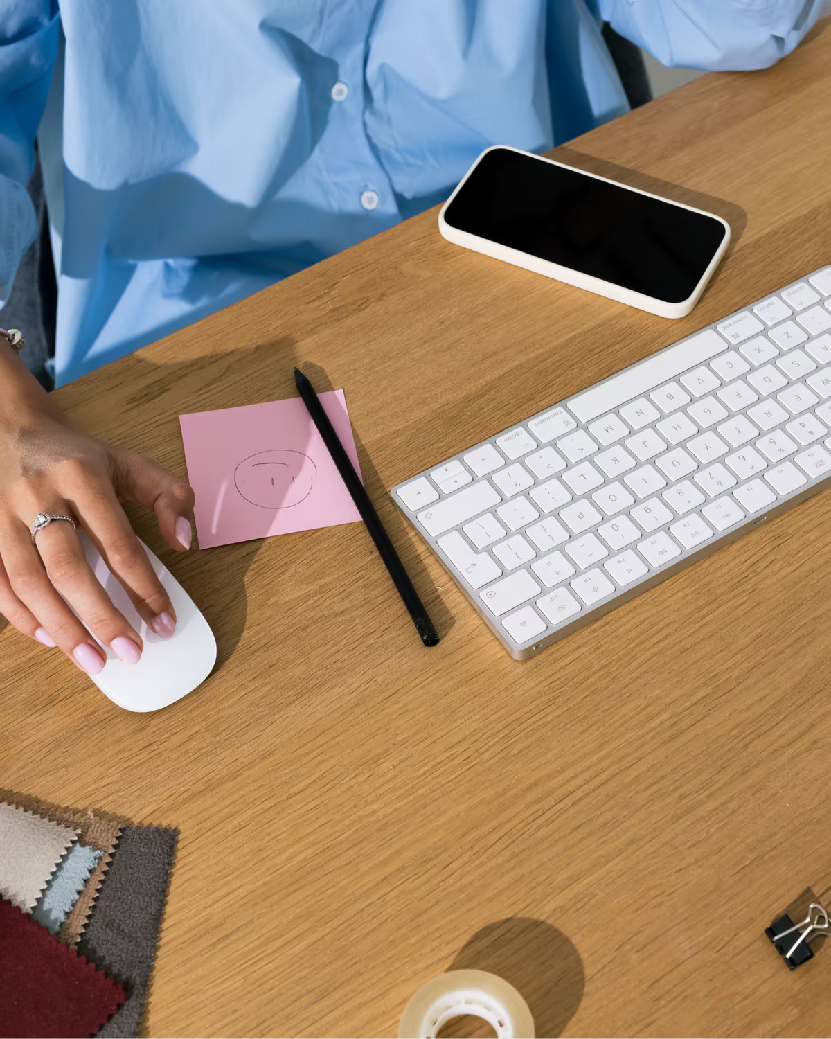 Hände an Maus und Tastatur auf einem Holzschreibtisch, daneben Smartphone, Notizzettel und Stoffmuster