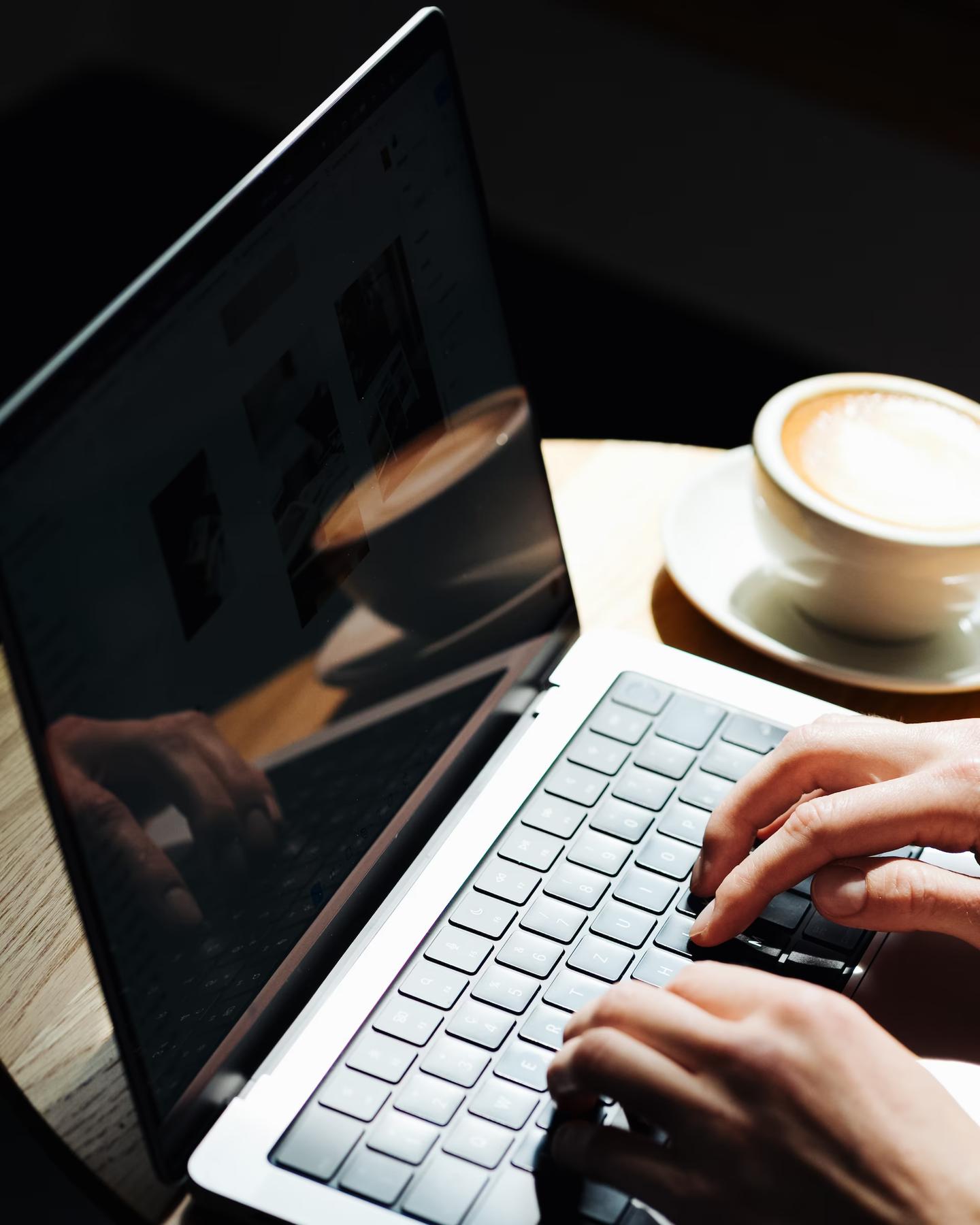 Hände tippen auf einem Laptop, daneben ein Cappuccino