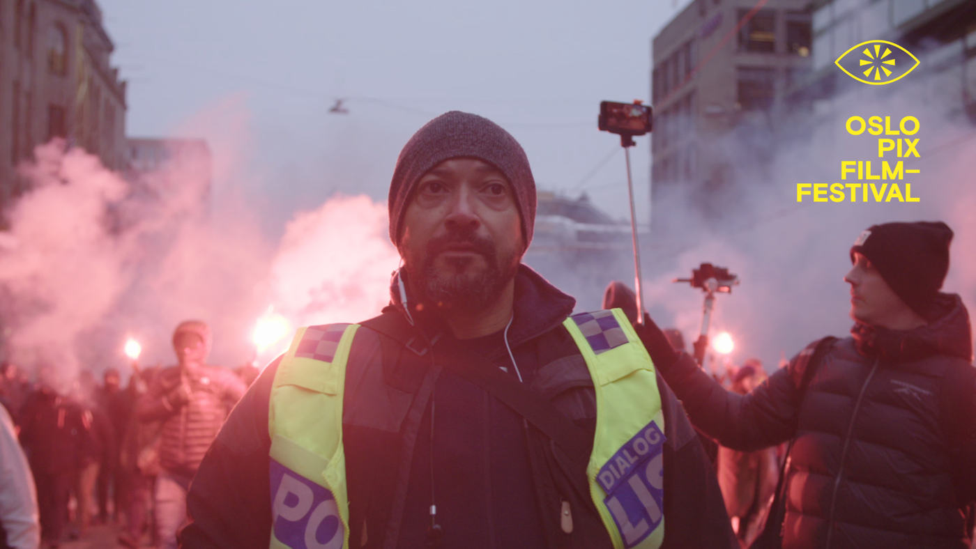 En politimann i gul vest med teksten «Dialog» står midt i en demonstrasjon med røyk og fakler, omgitt av mennesker og mobilkameraer.