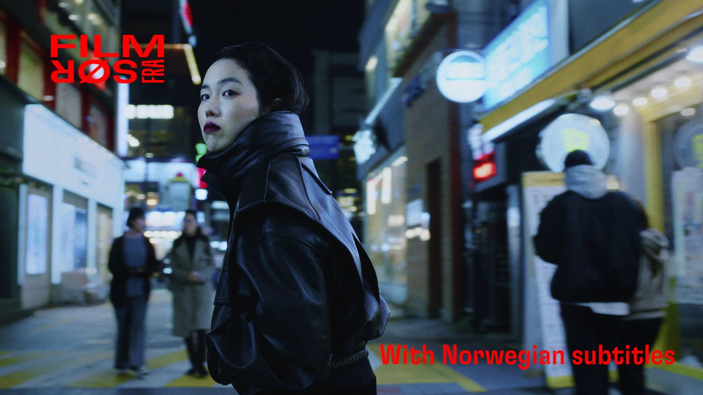 En kvinne i mørk skinnjakke snur seg og ser bakover mens hun går gjennom en opplyst bygate om natten.