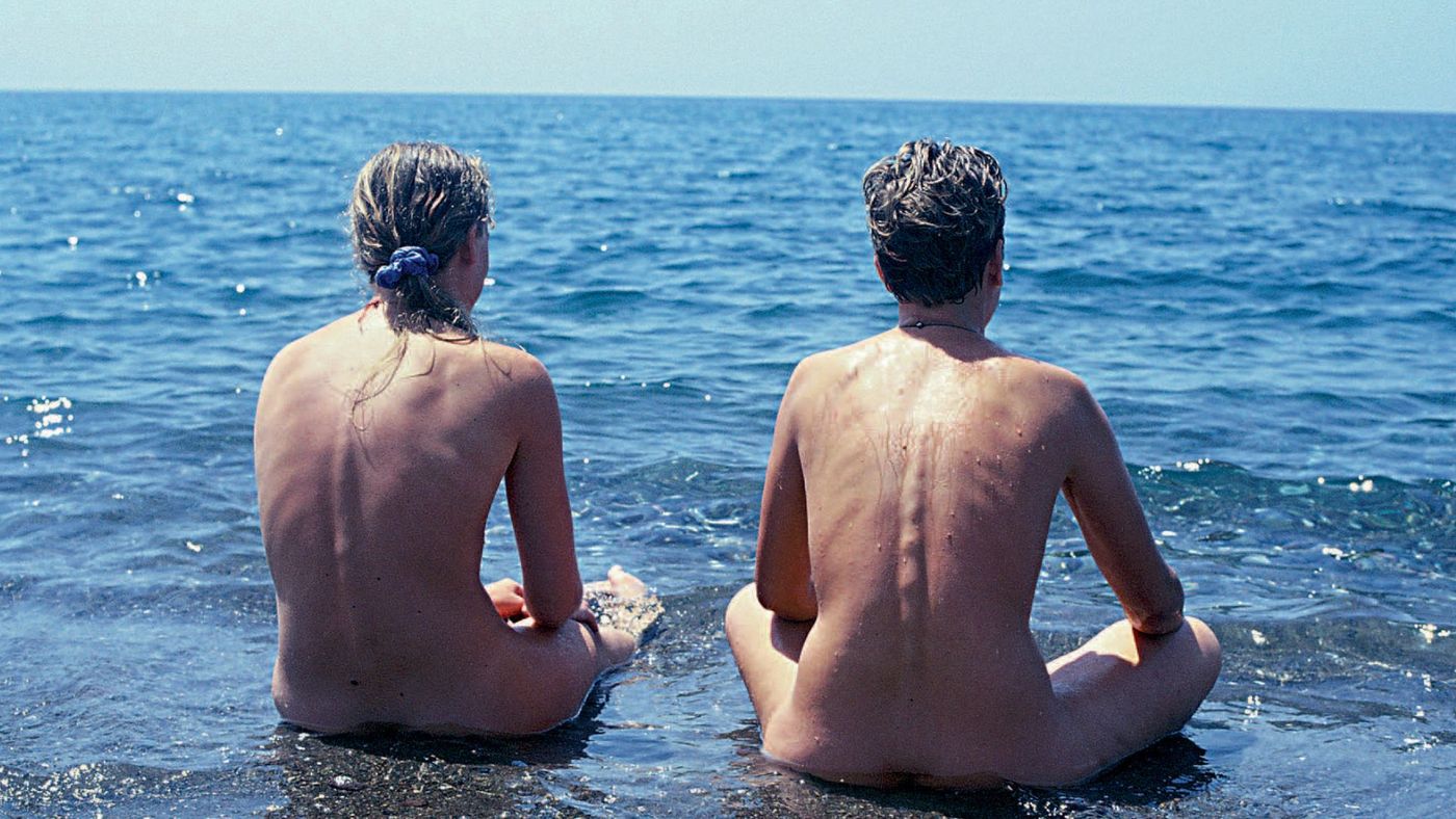 Bildet viser to nakne kvinner som sitter på strandkanten med bena i kryss og ser utover havet. Himmelen er skyfri. Vannet er noe bølgede. Hun til venstre har lyst hår i hestehale og blå hårstrikk. Hun andre har kort mørkt hår og et smykke på.