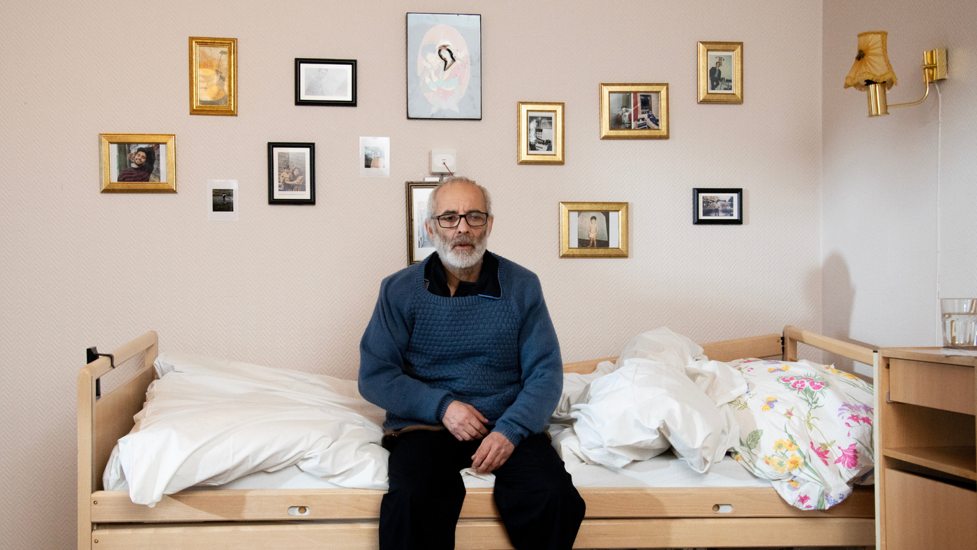 En eldre mann sitter på en seng. På veggen bak henger det bilder i rammer. Det gir inntrykk av å være på et sykehjem eller eldrehjem.
