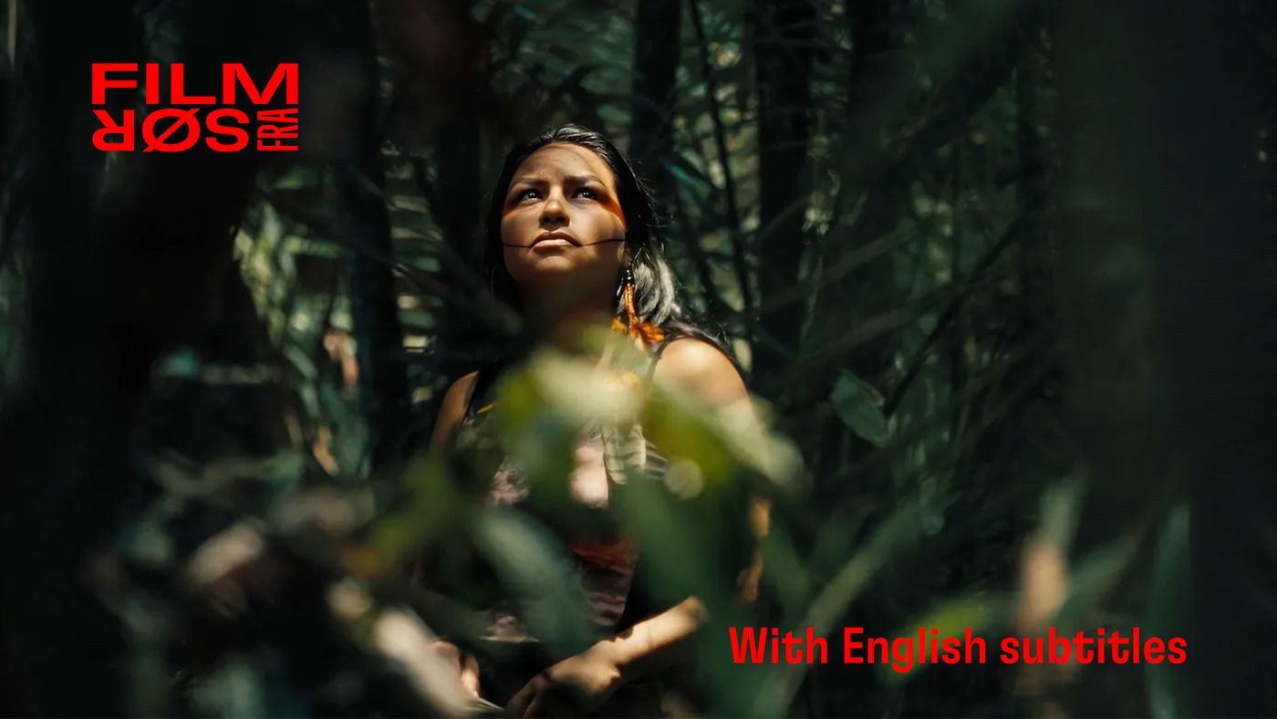 En kvinne står omgitt av grønne planter i skogen, badet i sollys som filtreres gjennom bladene.